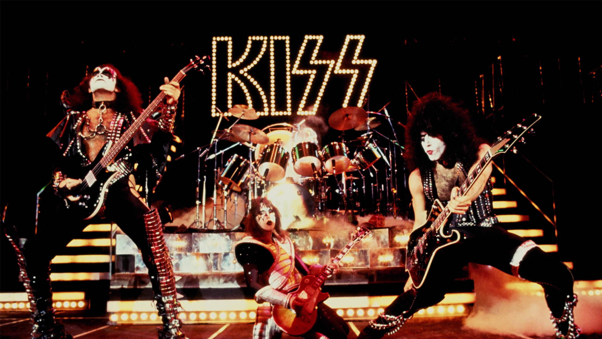 Kiss樂隊現場演唱會 壁紙 — KISS樂隊成員以全妝和舞台服裝在明亮燈光下表演，身後有發光的KISS標誌。