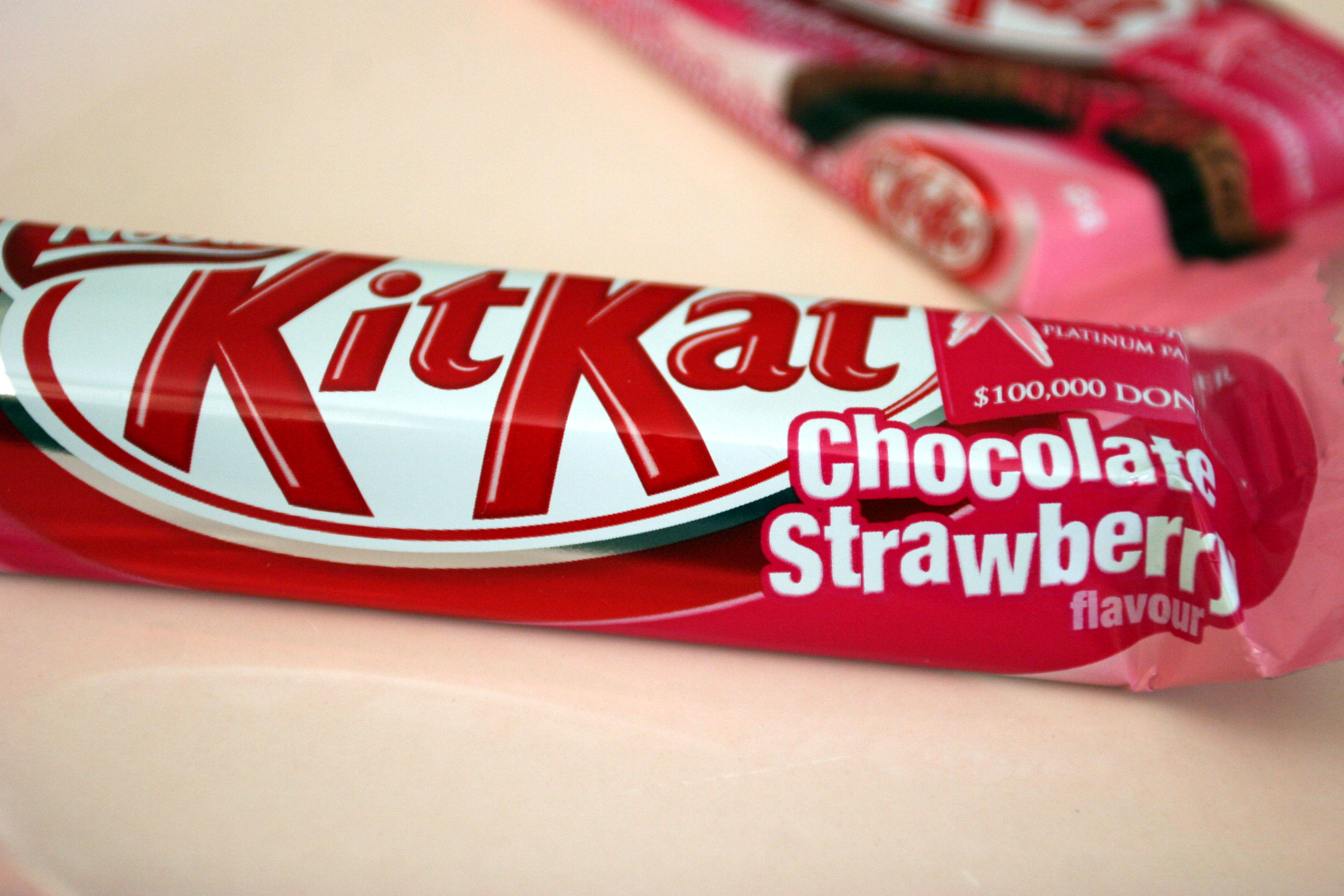 Kit Kat 巧克力草莓口味 壁紙 — 紅色和白色KitKat朱古力草莓味棒的近距離特寫，背景有模糊的粉紅色包裝。