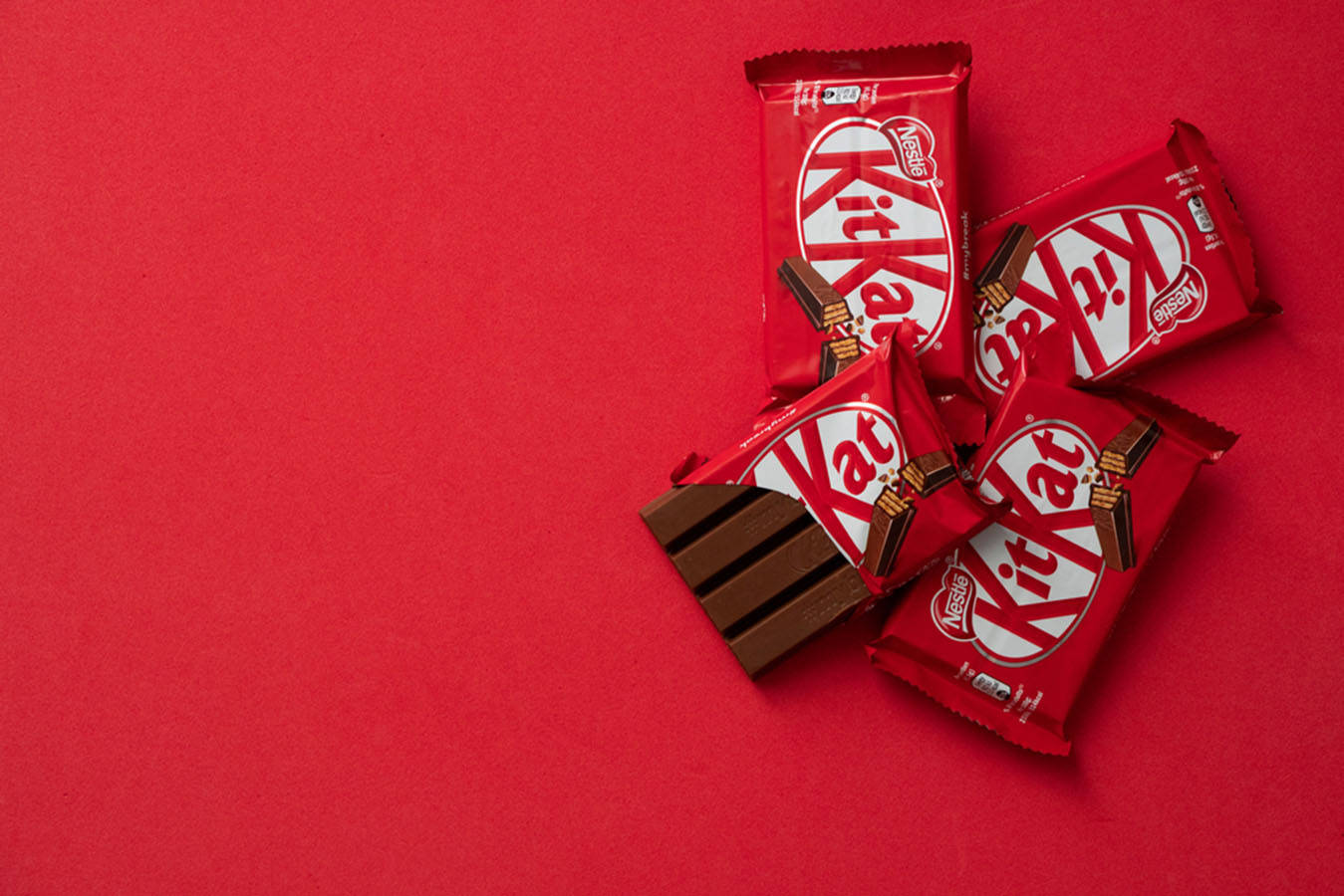 KitKat 紅色背景 壁紙 — 五條紅色包裝的KitKat朱古力棒散落在純紅色背景上，其中一條已拆開包裝，露出分段的朱古力。