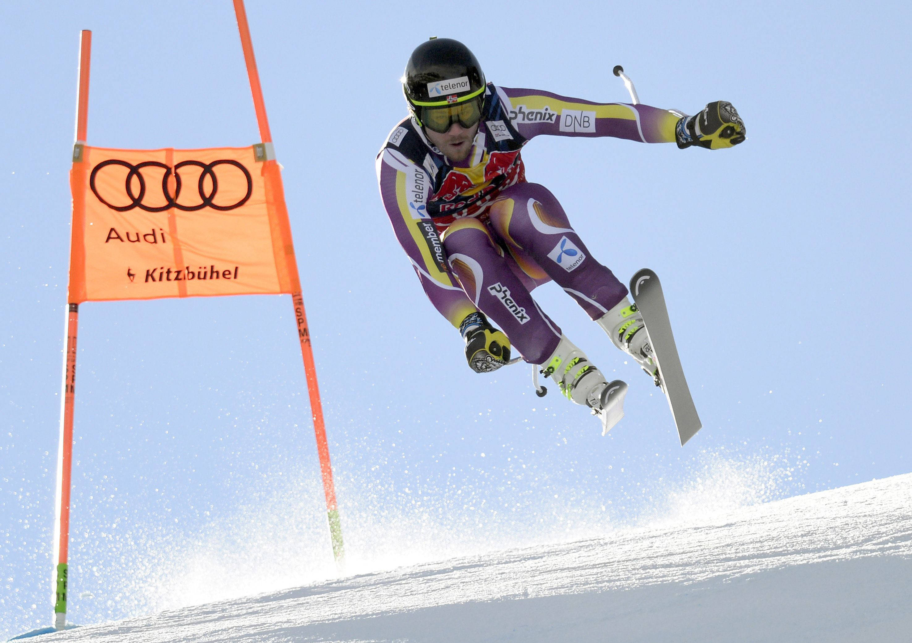 Kjetil Jansrud 領先高山滑雪 壁紙 — 一名滑雪運動員在白雪覆蓋的斜坡上空飛過一個橙色 Audi 閘門，背景是清澈的藍天。
