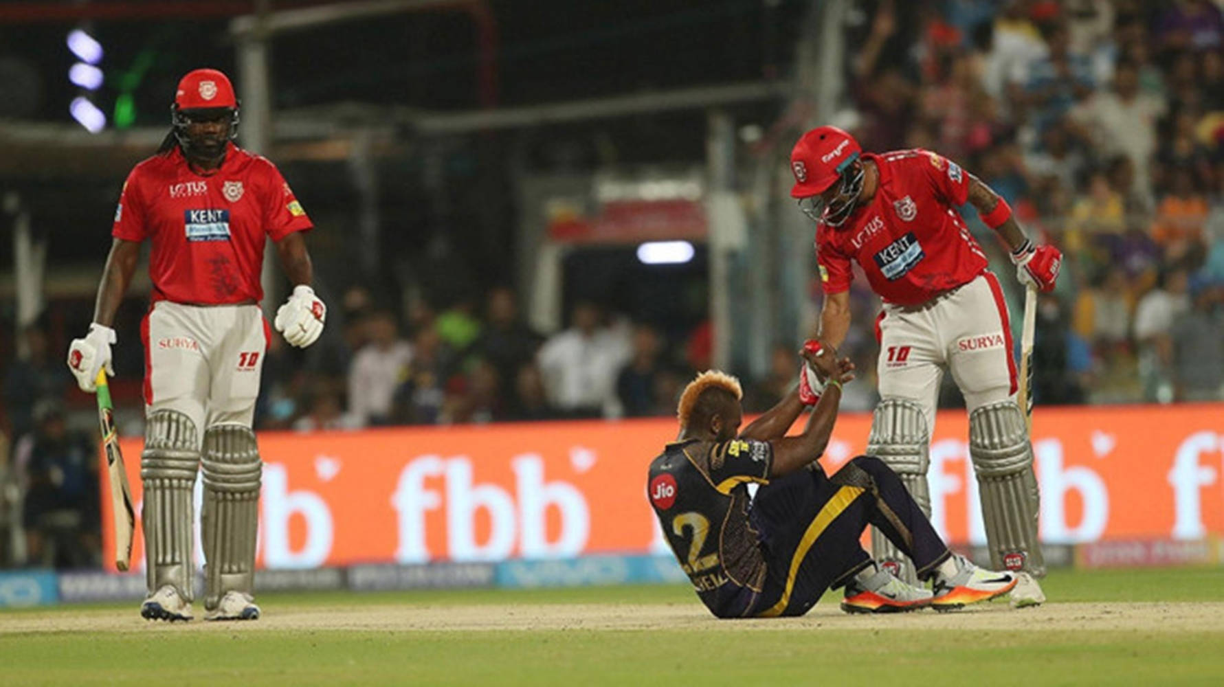KL拉胡爾對戰 Kolkata Knight Riders 壁紙 — 兩名身穿紅色球衣的板球運動員在燈光明亮的體育場場地上，協助一名身穿海軍藍裝備的倒地隊友。