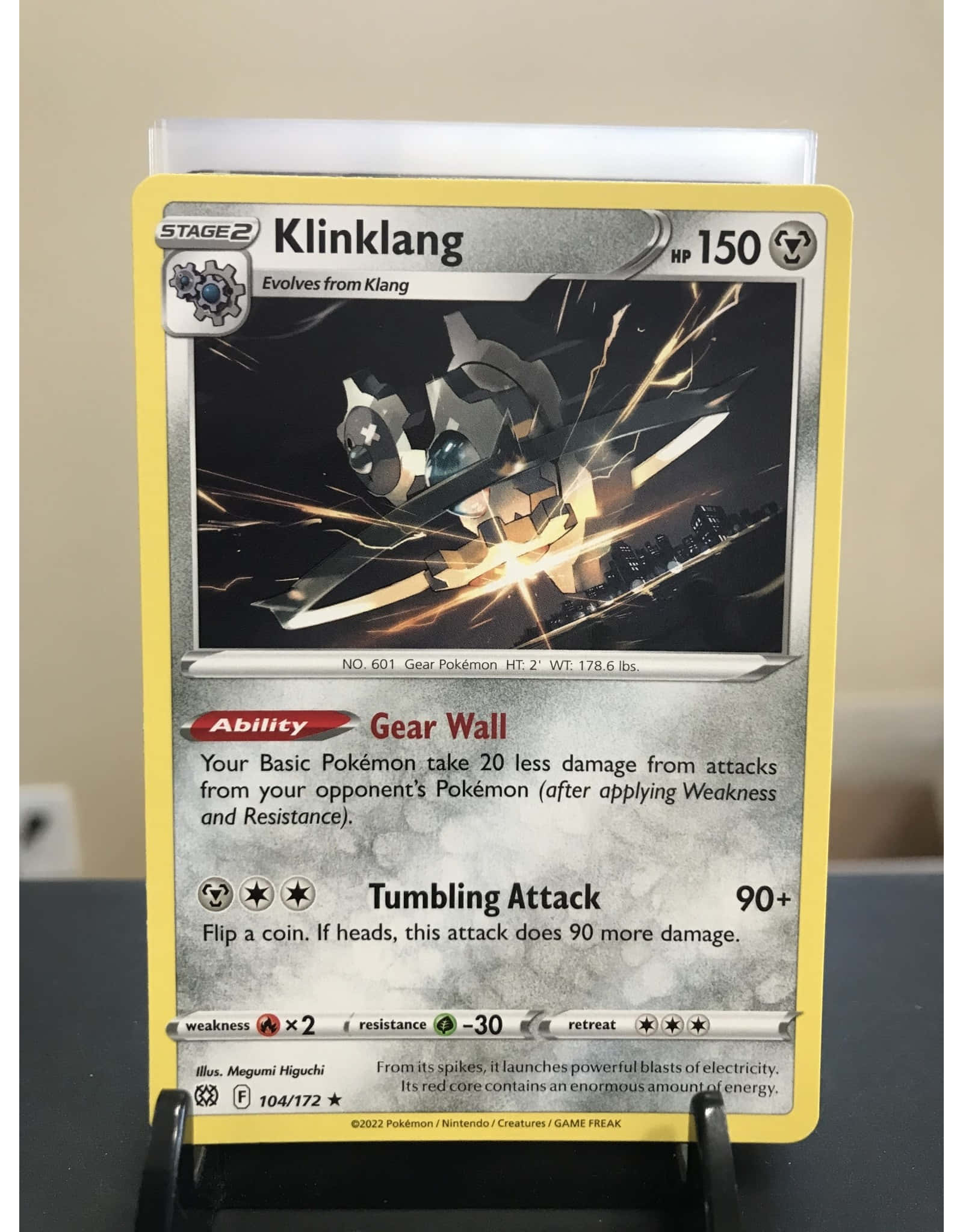 Klinklang 寵物小精靈卡牌立於支架上 壁紙 — 一張黃色邊框的寶可夢卡，描繪了齒輪怪，帶有金屬機械設計和發光能量效果。