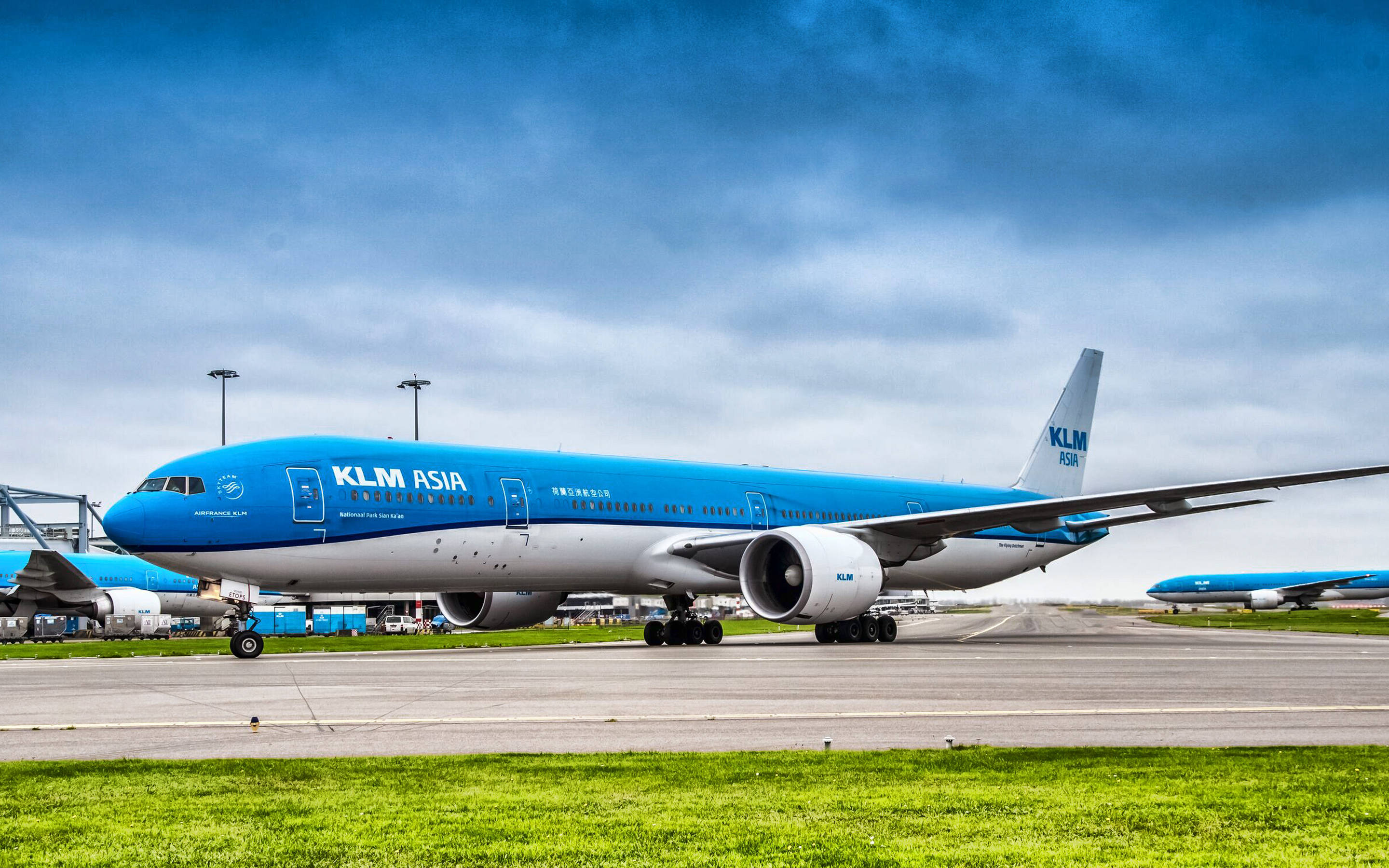 KLM Asia 客機機場靜態壁紙 壁紙 — 一架 KLM Asia 波音 777 停泊在機場停機坪上，背景是戲劇性的多雲天空，前景是充滿活力的綠色草地。