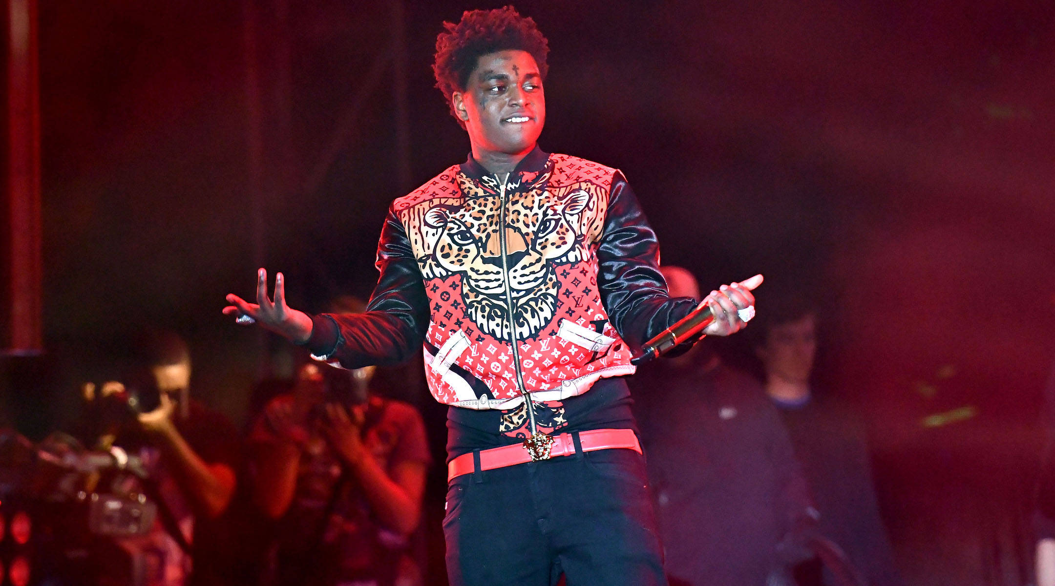Kodak Black演唱會瞬間 壁紙 — 一位身穿豹紋外套的年輕男表演者在昏暗的舞台上唱歌，舞台沐浴在紅光中。