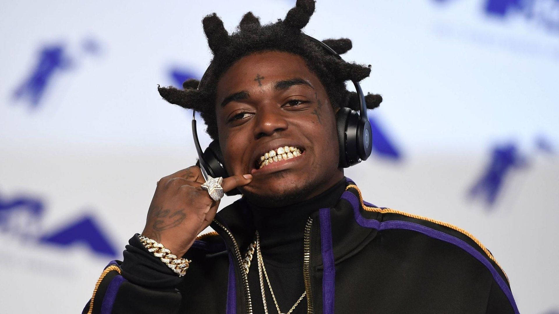 Kodak Black 星形戒指 壁紙 — 一位微笑的男士，留著辮子頭，戴著耳機，穿著飾有金鏈的黑色和紫色外套，正在調整麥克風。