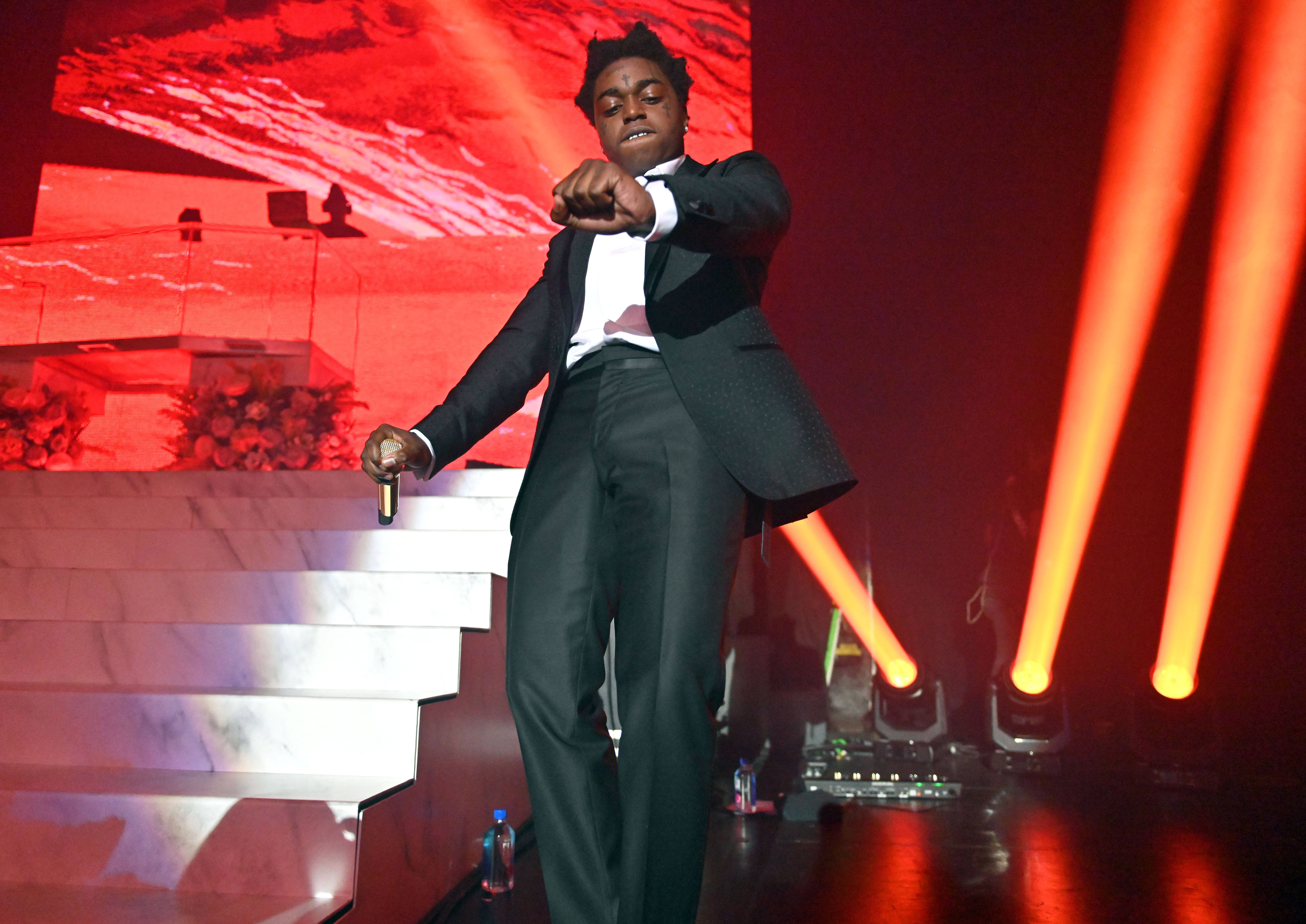 Kodak Black 西裝造型 壁紙 — 一位身穿黑色西裝的表演者主宰著一個沐浴在戲劇性紅光和發光光束中的舞台。