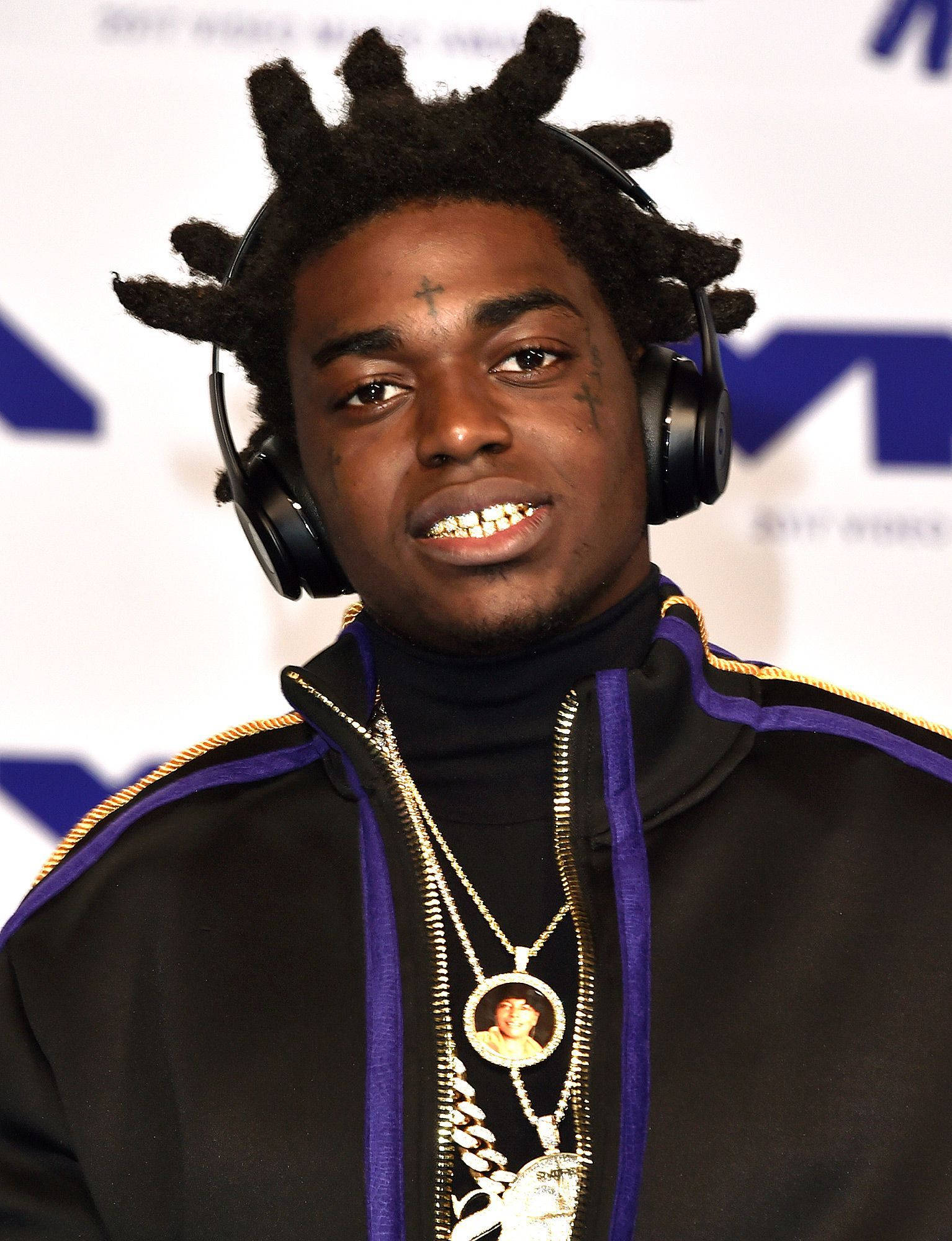微笑的 Kodak Black 壁紙 — 一位年輕男子留著辮子頭和金色牙齒，戴著耳機和多層項鍊，背景為品牌背景。