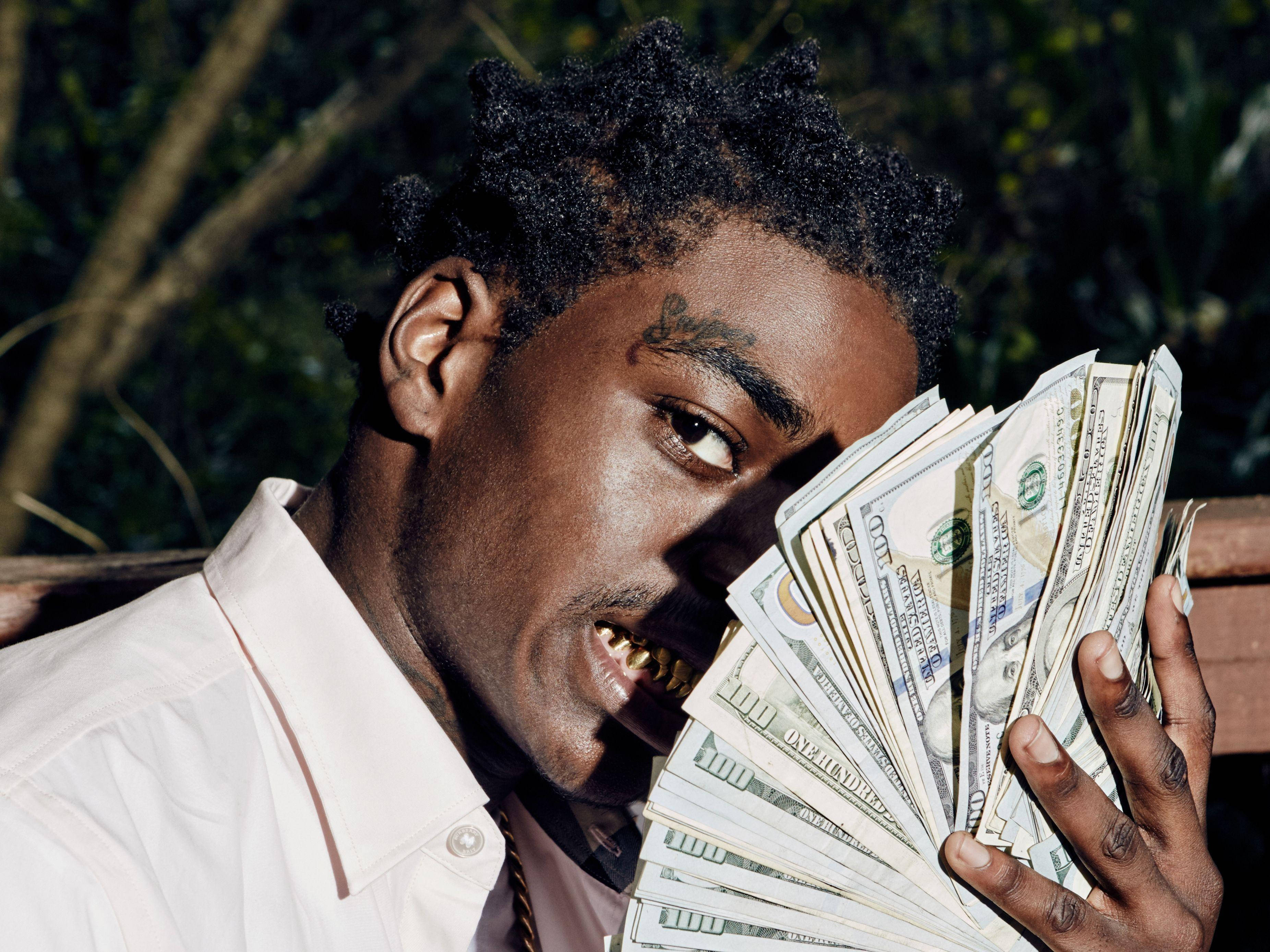 Kodak Black 展現財富 壁紙 — 一個有金牙和紋身的男人，在模糊的綠色背景下，拿著一疊百元美鈔扇子，專注地看著鏡頭。