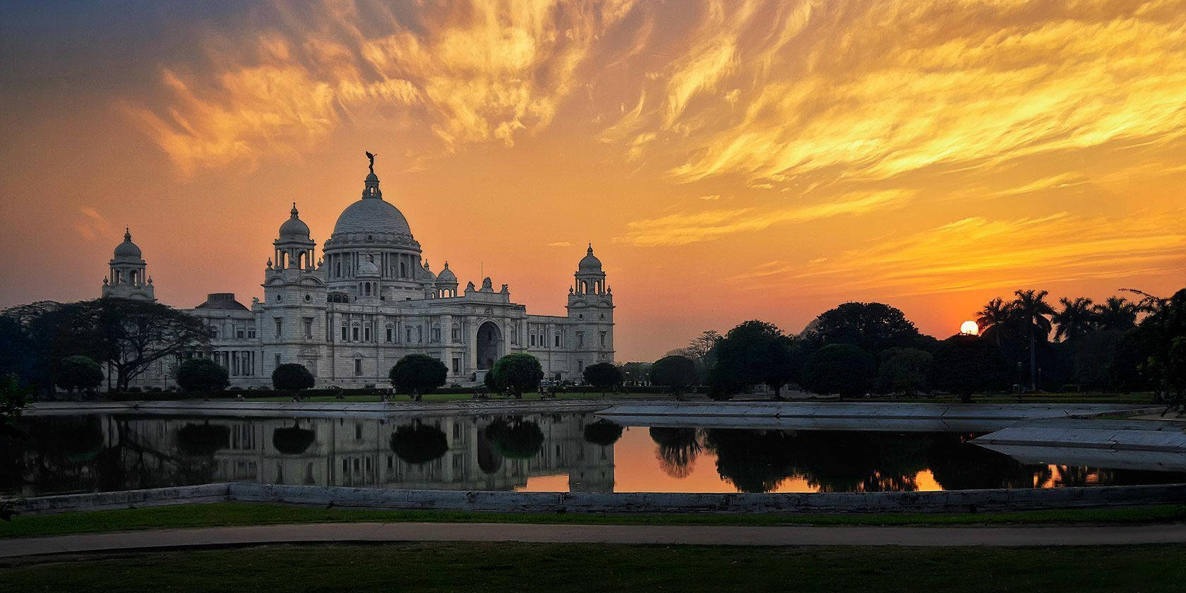 Kolkata 日落天空 壁紙 — 一座宏偉的白色新古典主義建築在平靜的水面上倒映，天空是火熱的橙黃色日落。