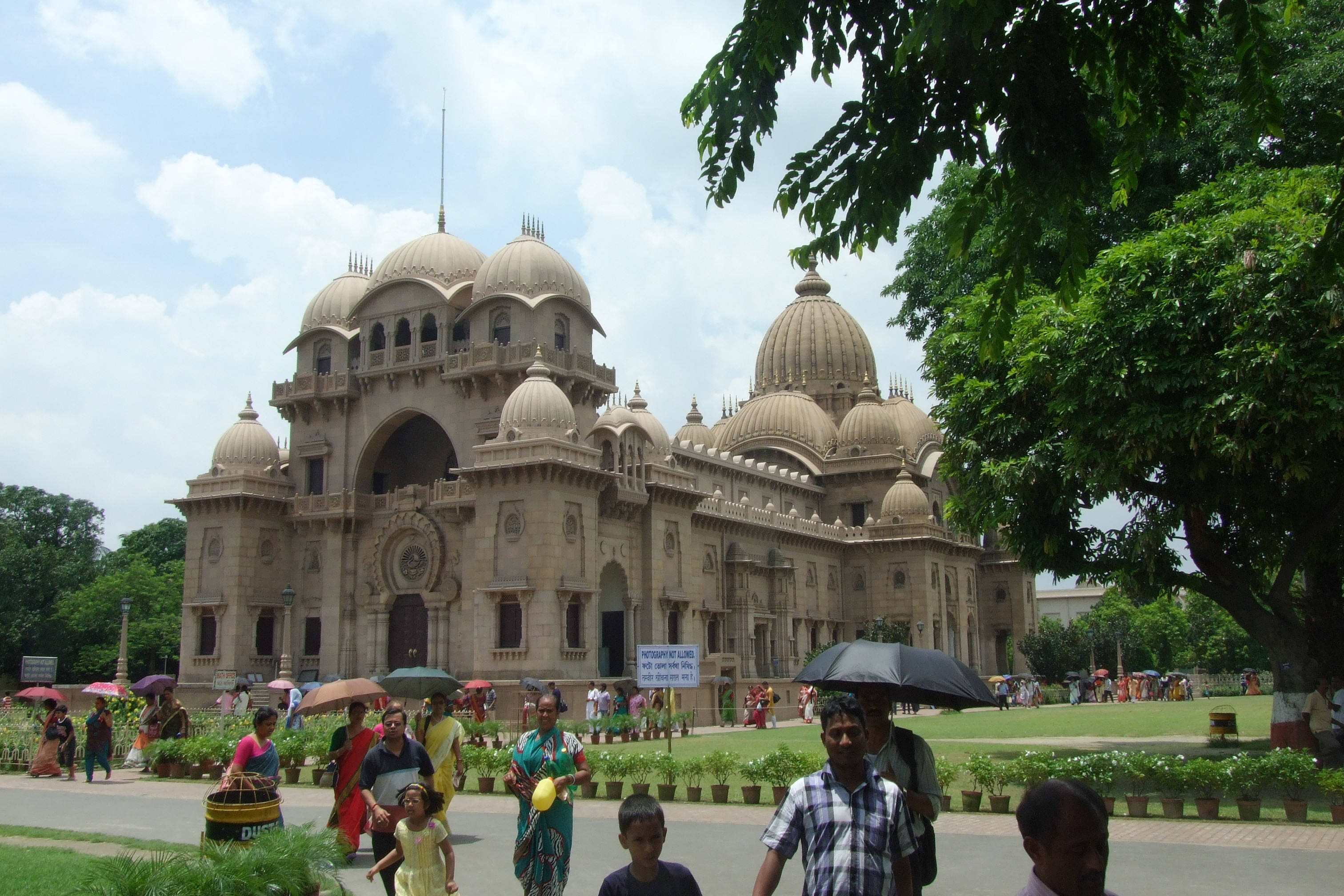 Kolkata 貝魯爾馬特 壁紙 — 巨大的米色圓頂寺廟帶有精緻雕刻，在明亮的天空下矗立，遊客撐著傘在鬱鬱蔥蔥的綠色公園中漫步。