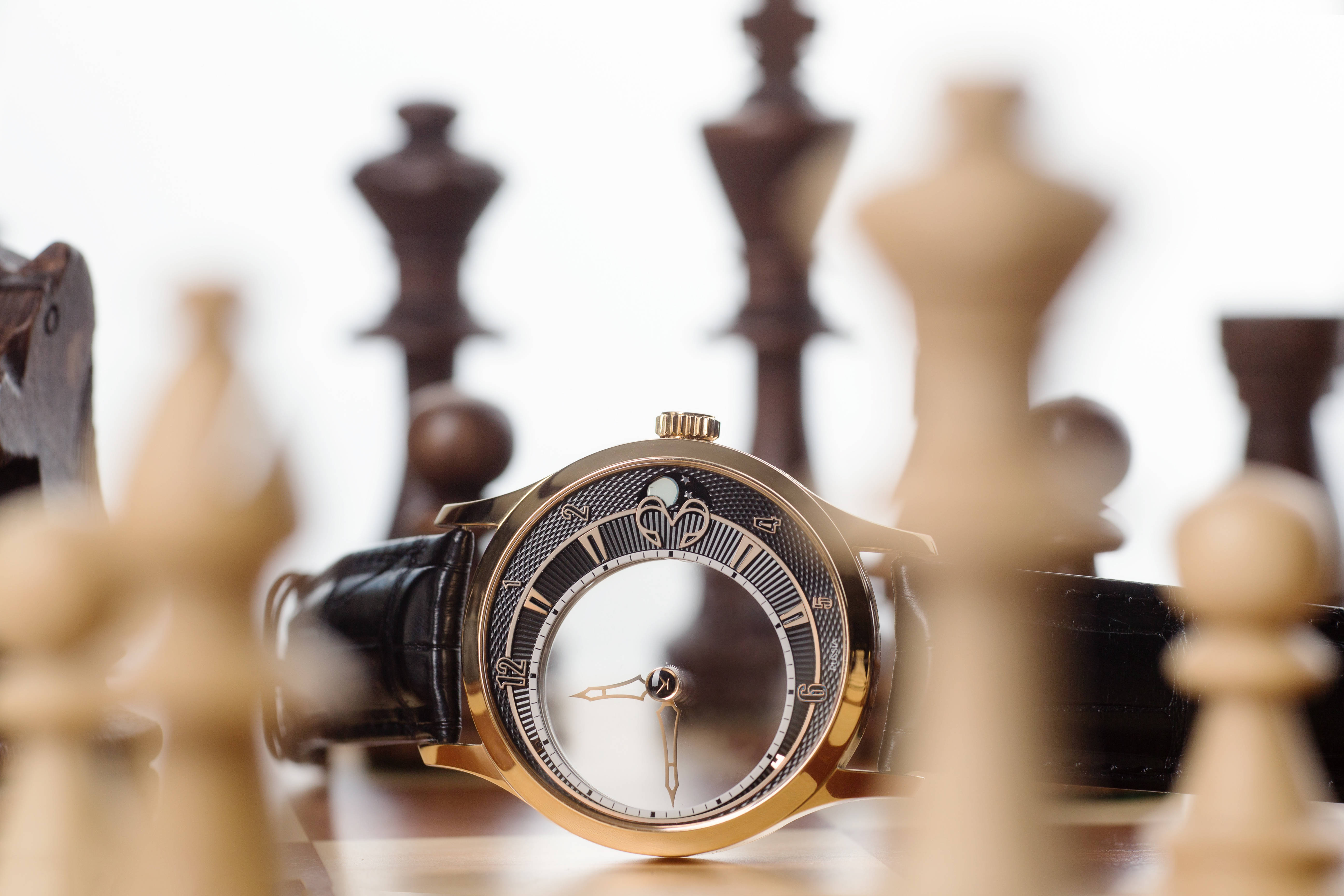 Konstantin Chaykin 透明腕錶 壁紙 — 一隻金色手錶配黑色皮帶，放在一個被模糊木製棋子包圍的棋盤上。