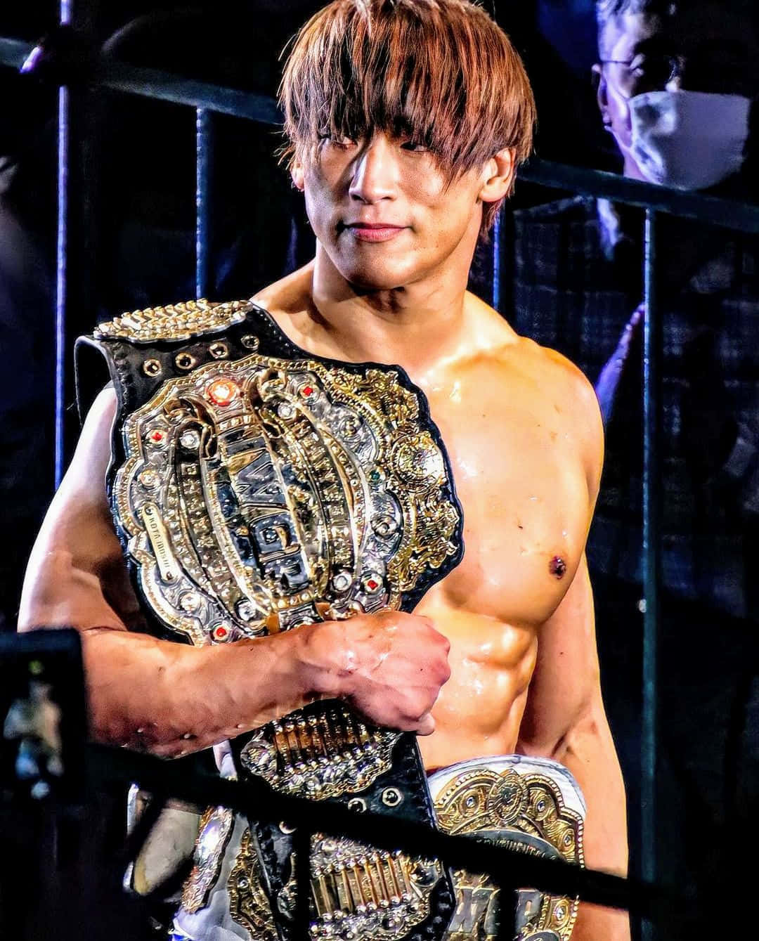 Kota Ibushi 舉起IWGP洲際冠軍腰帶 壁紙 — 一個肌肉發達的摔角手，皮膚濕潤，棕色頭髮，在昏暗的擂台上拿著閃閃發光的冠軍腰帶。