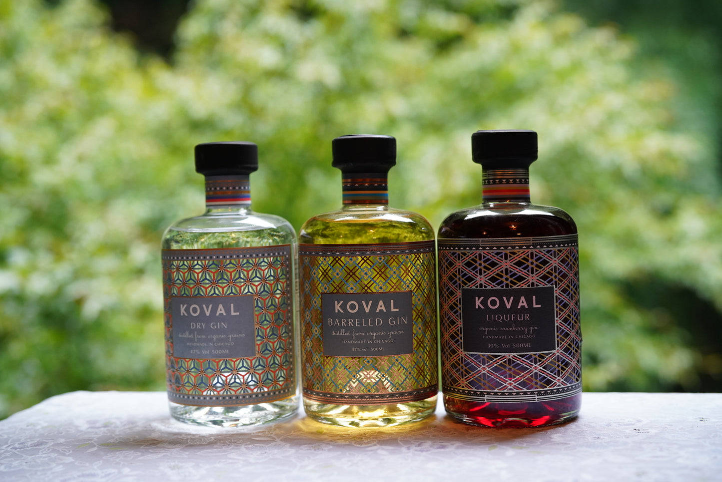 三支Koval烈酒瓶，帶有彩色標籤，放在白色表面上，背景有綠色樹葉。