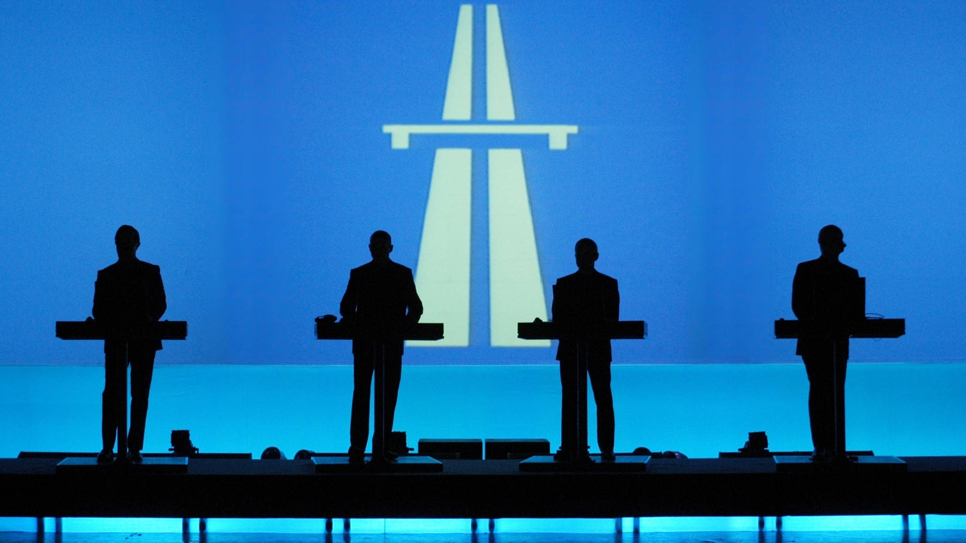 Kraftwerk《Autobahn》專輯封面 壁紙 — 四個剪影音樂家站在鍵盤前，舞台上籠罩著冷藍色燈光，背後有發光的高速公路標誌投影。
