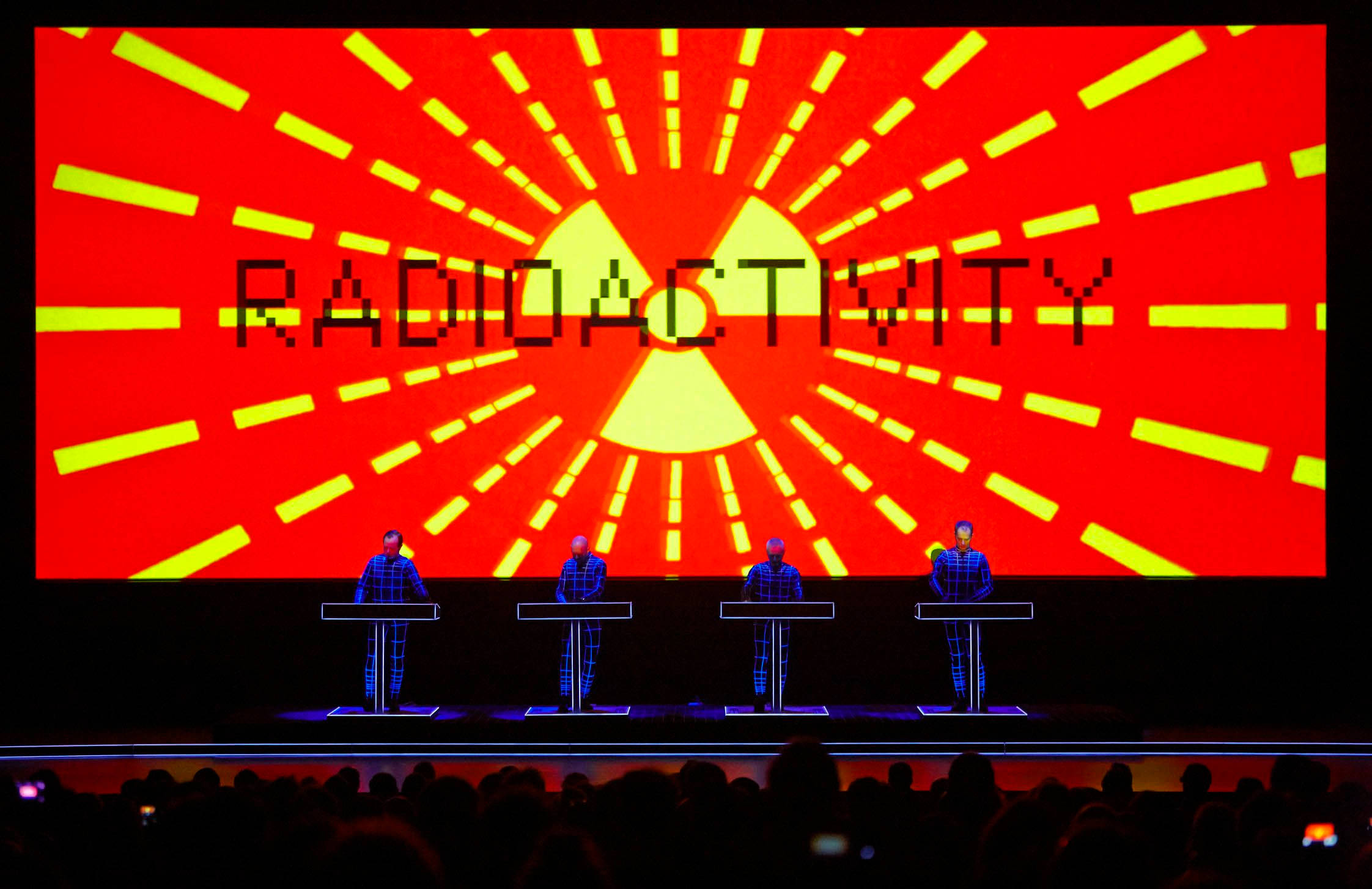 Kraftwerk《Radioactivity》塗鴉藝術 壁紙 — 四位身穿格仔西裝的音樂家在舞台上表演，舞台上方有一個巨大的紅色屏幕，顯示「RADIOACTIVITY」和一個發光的黃色輻射標誌。