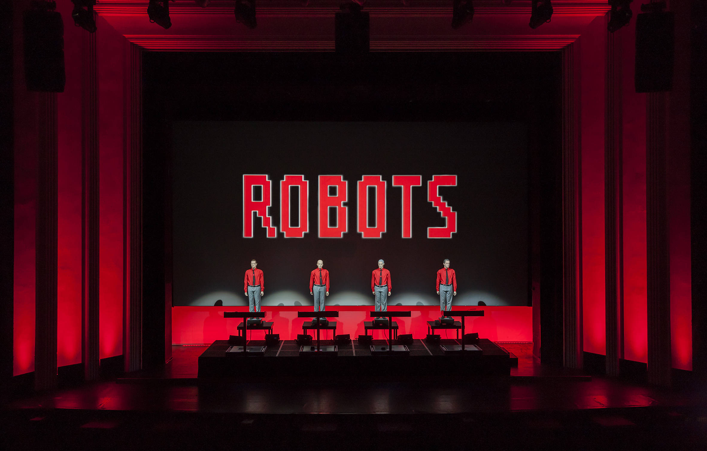 Kraftwerk《Robots》現場舞台表演 壁紙 — 四位身穿紅色外套的表演者站在昏暗的舞台上，紅色燈光下，身後有一個大型像素化的「ROBOTS」標誌。