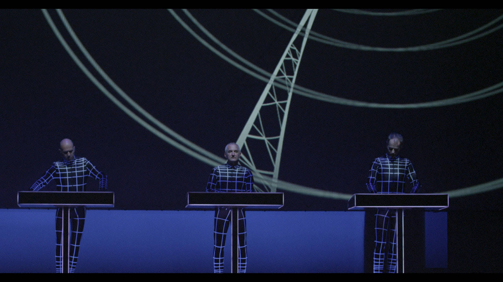 Kraftwerk《Trans-Europe Express》演唱會 壁紙 — 三個光頭人物穿著發光藍色網格套裝，站在講台上，背景黑暗，有彎曲的霓虹線條和塔樓輪廓。