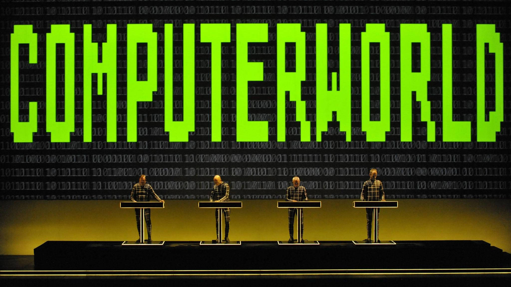Kraftwerk 在 Computer World 巡迴演出中的現場表演 壁紙 — 四個穿著格紋西裝的人物站在講台上，背景是滾動的二進制代碼，上方有發光的綠色「COMPUTERWORLD」文字。