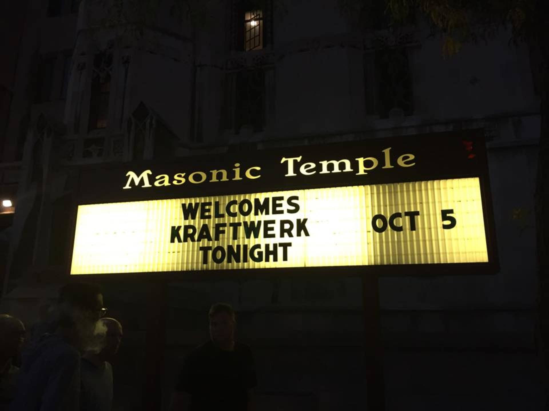 Kraftwerk 在共濟會堂演出 壁紙 — 夜晚發光的招牌宣佈 Kraftwerk 於10月5日在共濟會神廟的音樂會，下方有剪影人物。