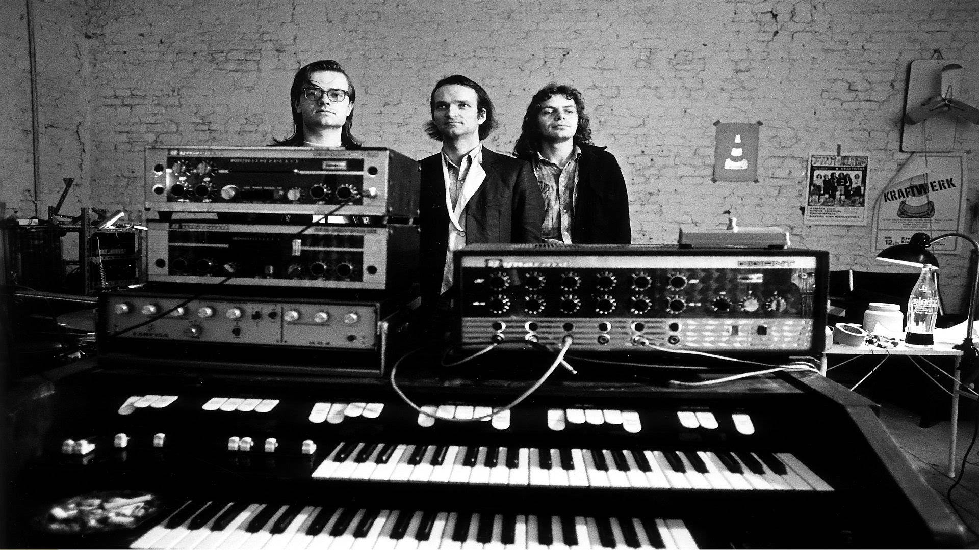 Kraftwerk 在錄音室創作 壁紙 — 三個男人站在復古合成器和鍵盤後面，工作室雜亂，有裸露的磚牆和海報。
