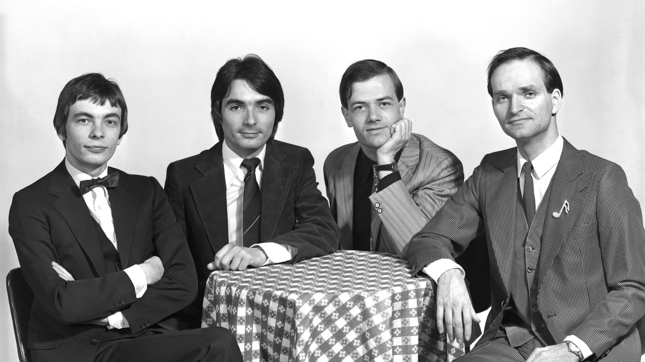 Kraftwerk 德國樂團黑白版 壁紙 — 四位身穿西裝的男士在格仔枱布周圍正式擺姿勢，經典黑白影樓肖像。