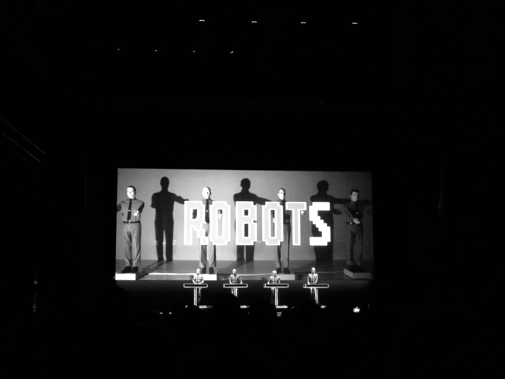 Kraftwerk — 機器人主題現場演出 壁紙 — 黑白舞台表演，演員穿著機械人服裝，在明亮燈光下拼出 ROBOTS。