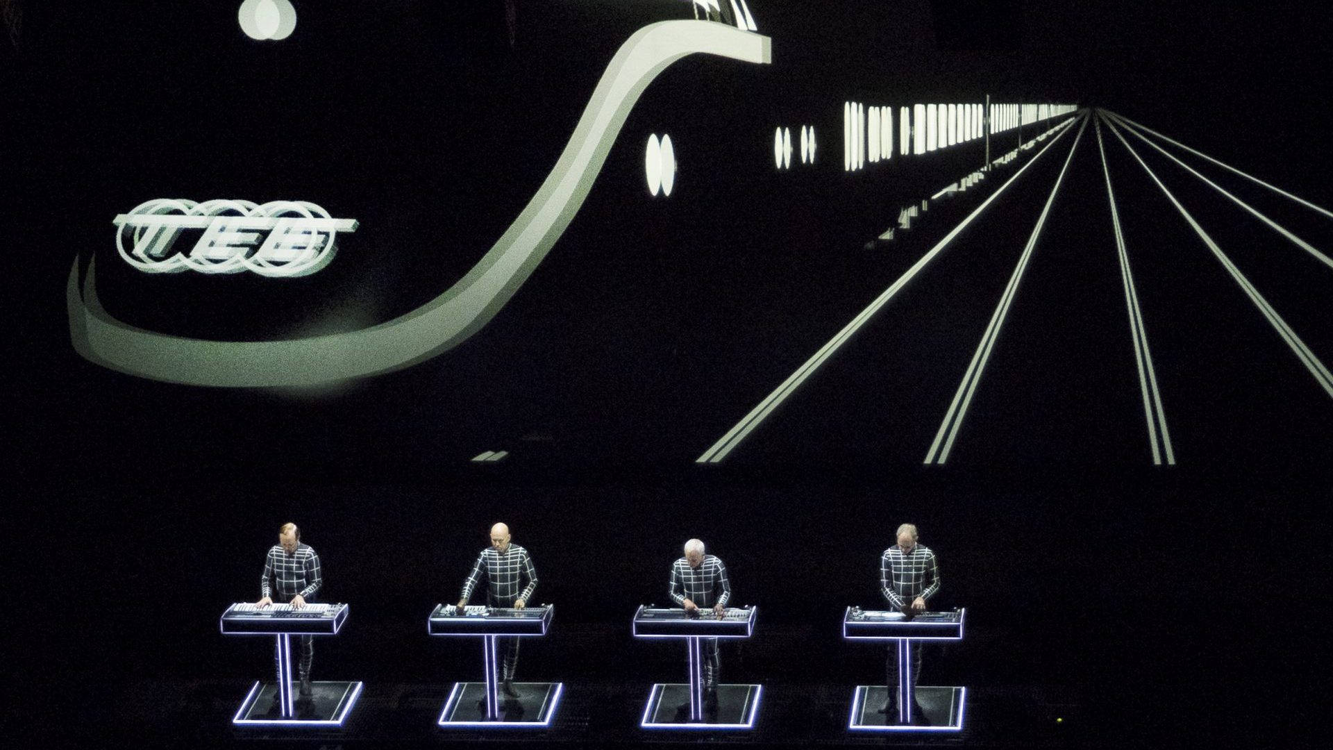Kraftwerk 高速公路現場演出 壁紙 — 四位身穿格仔恤衫嘅音樂家喺黑暗嘅音樂廳入面，喺一個大型抽象白色燈光裝置之下，喺發光嘅舞台上表演。