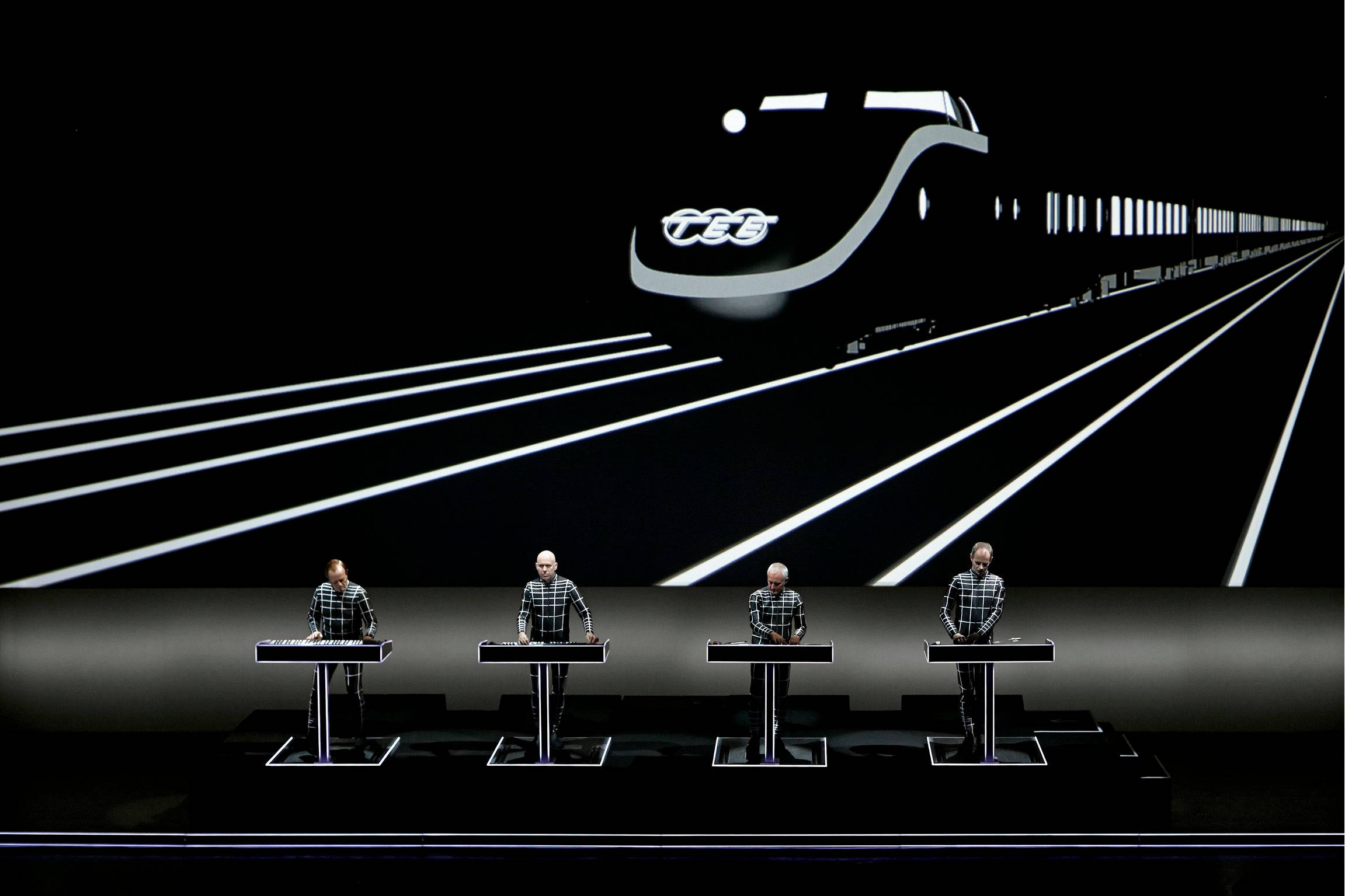Kraftwerk 高速列車舞台背景 壁紙 — 四位身穿格仔恤衫的音樂家在黑暗的舞台上，在閃爍的未來火車投影下表演。