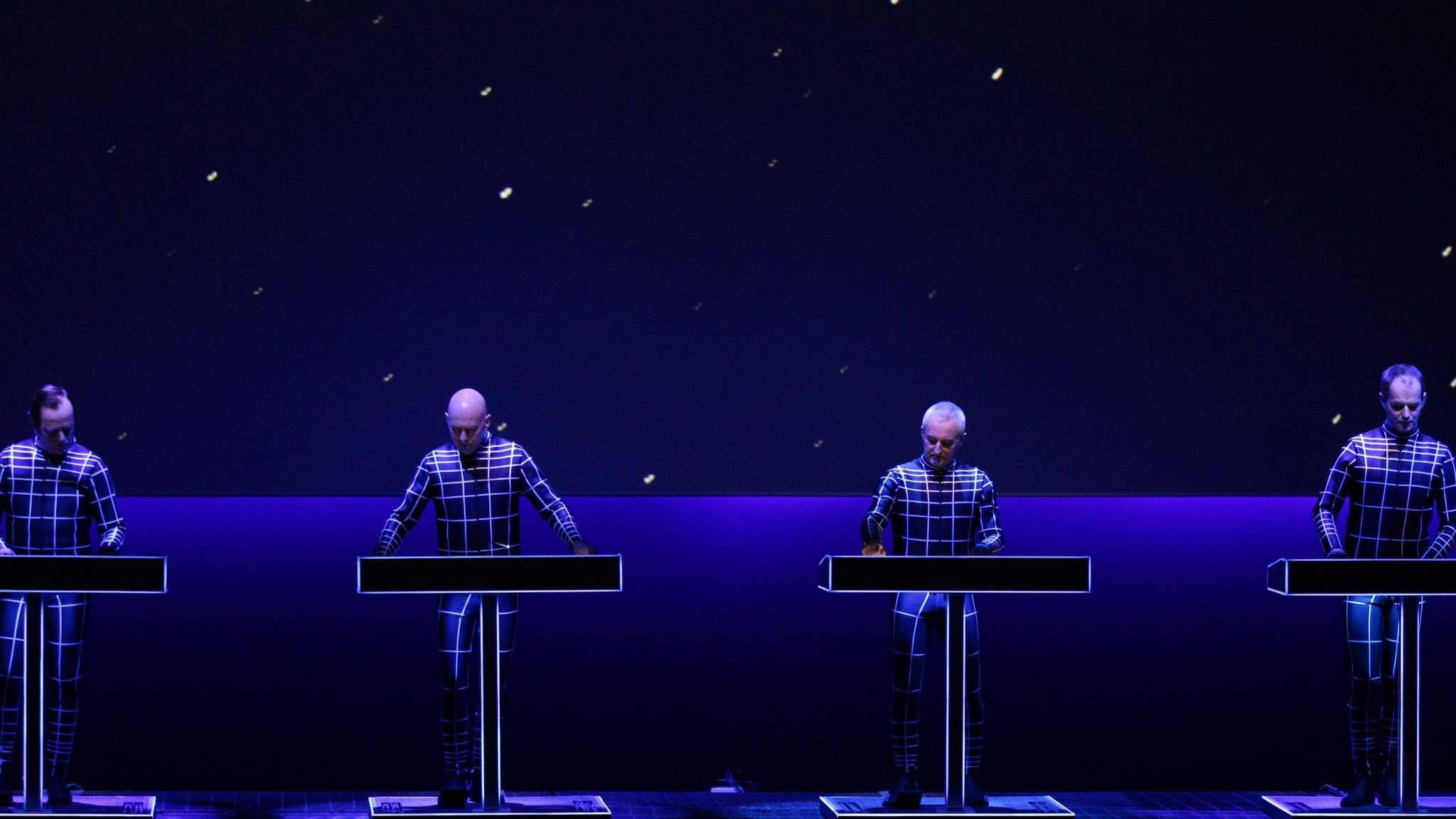 Kraftwerk《Trans-Europe Concert》星際背景 壁紙 — 四位光頭音樂家穿著格紋西裝，在深藍色舞台燈光下演奏鍵盤，伴隨著飄浮的火花。