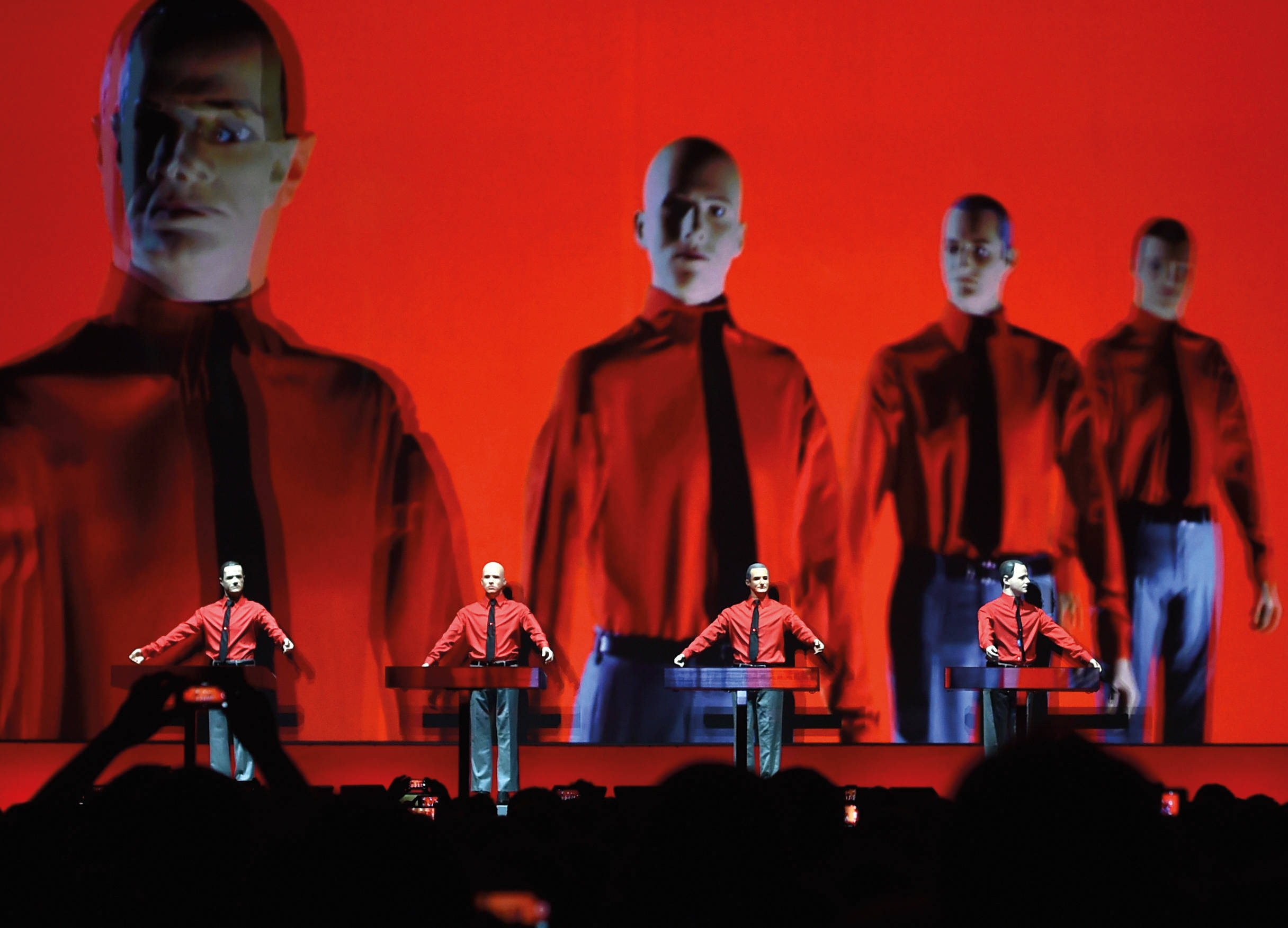 Kraftwerk紅衣現場演出 壁紙 — 四位身穿紅衫黑領帶的表演者在舞台上，在一個顯示他們面孔的巨大紅色屏幕下彈奏鍵盤。