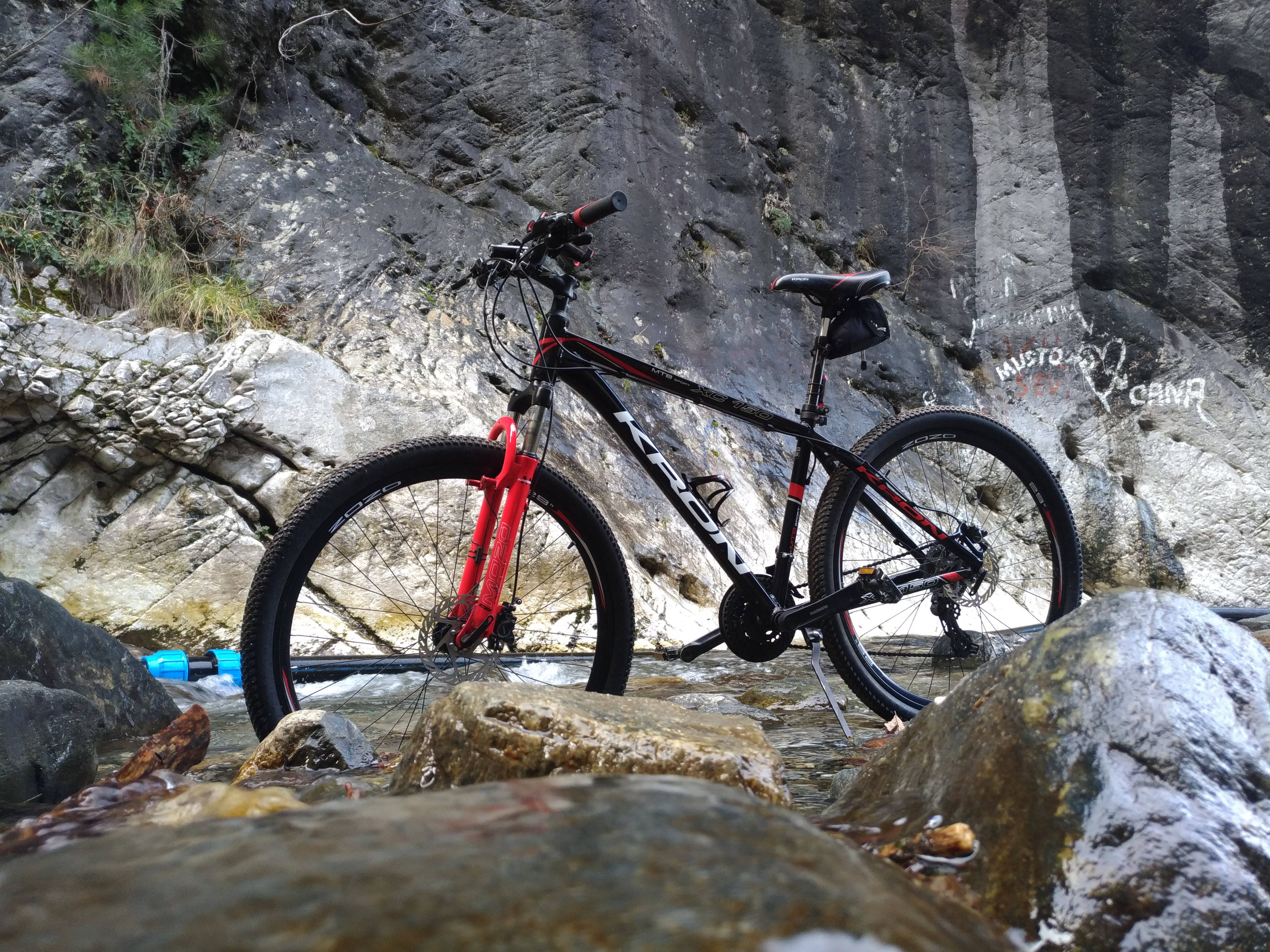 Kron山地自行車 壁紙 — 一架紅黑色山地單車佇立於淺山溪中，四周是濕石和崎嶇懸崖。