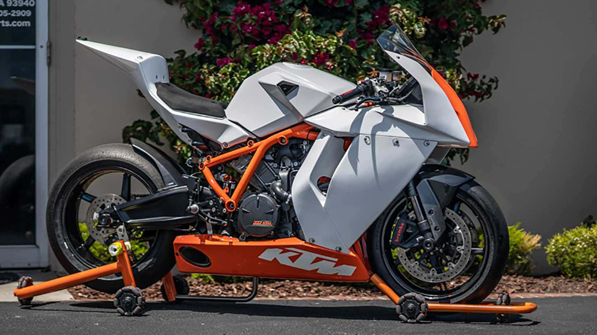 KTM 1190 RC8 圖片 壁紙 — 一架白色和橙色嘅KTM運動電單車喺支架上，停泊喺戶外，後面有開花嘅灌木叢。