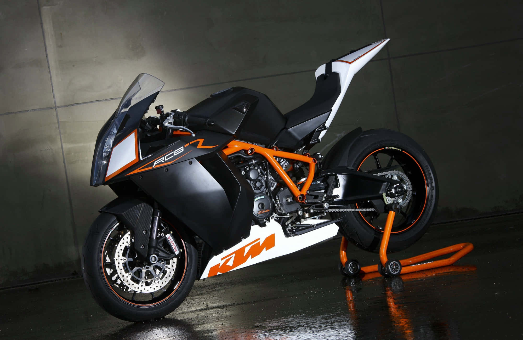 KTM 1190 RC8圖片 壁紙 — 一架時尚的黑色和橙色KTM RC8電單車停放在支架上，位於燈光昏暗的混凝土車庫中，地板濕潤反光。
