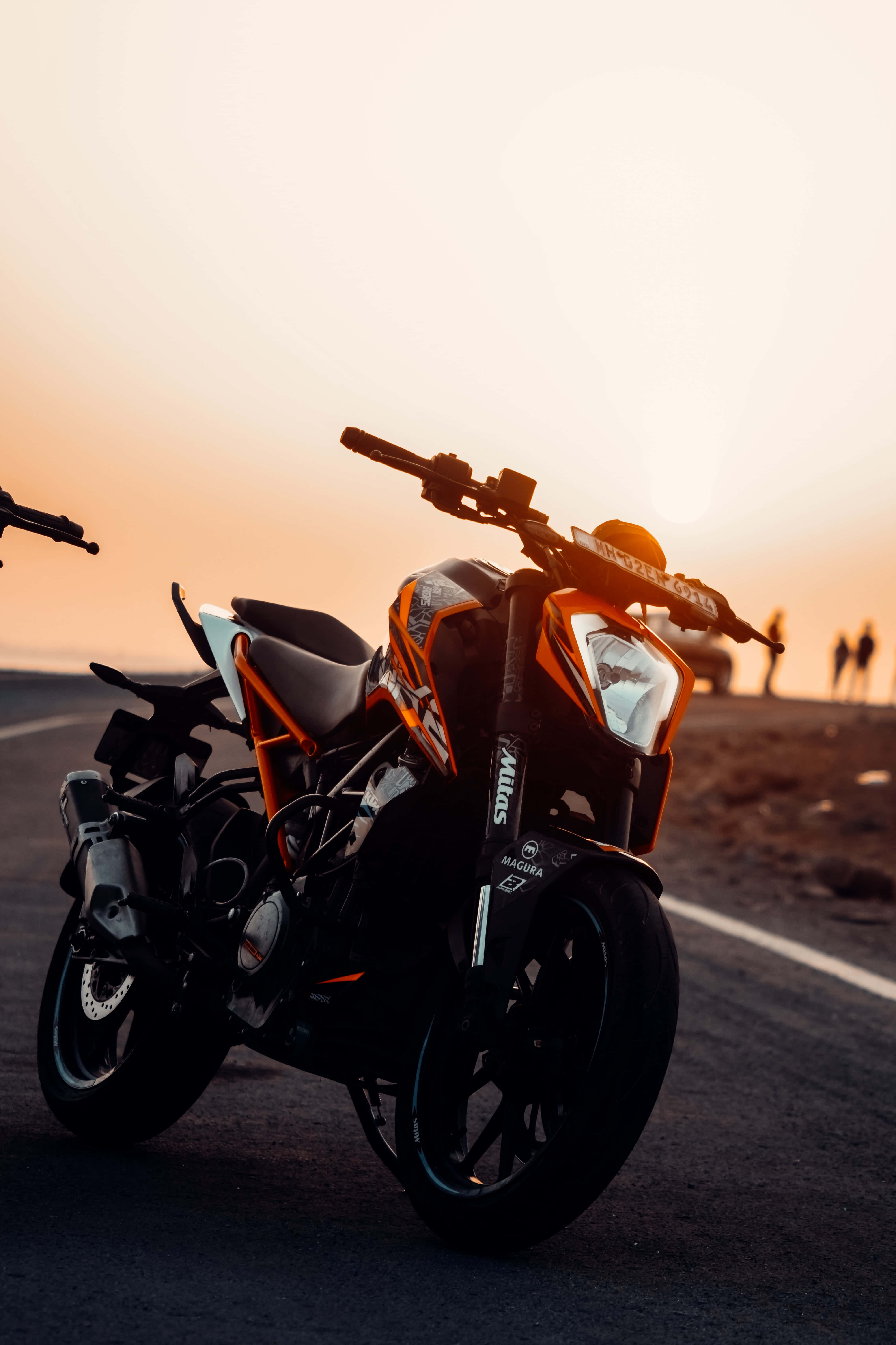 優雅KTM 390系列機車背景 壁紙 — 一輛時尚的橙色和黑色電單車在日落時停泊在空曠的道路上，遠處有剪影人物。