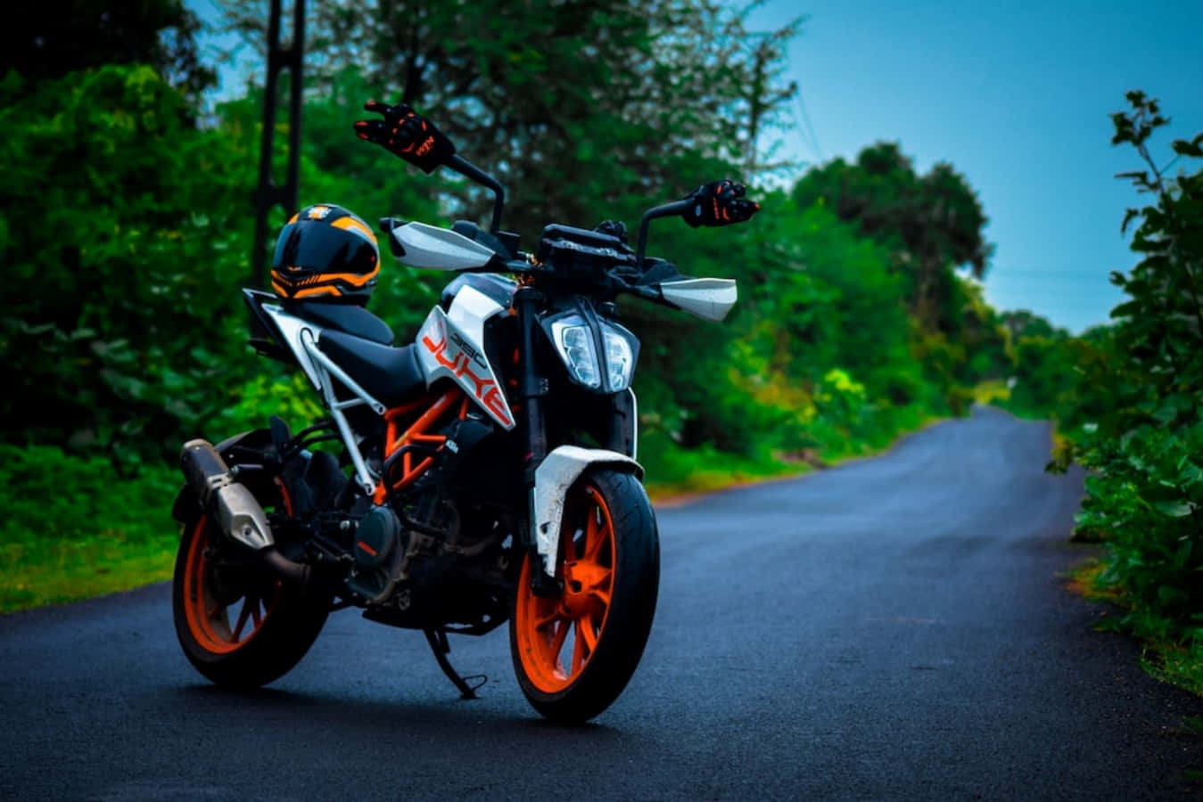 KTM 390照片 壁紙 — 一架橙色和白色的運動型電單車停泊在蜿蜒的林間道路上，周圍是茂密的綠樹，頭頂是清澈的藍天。