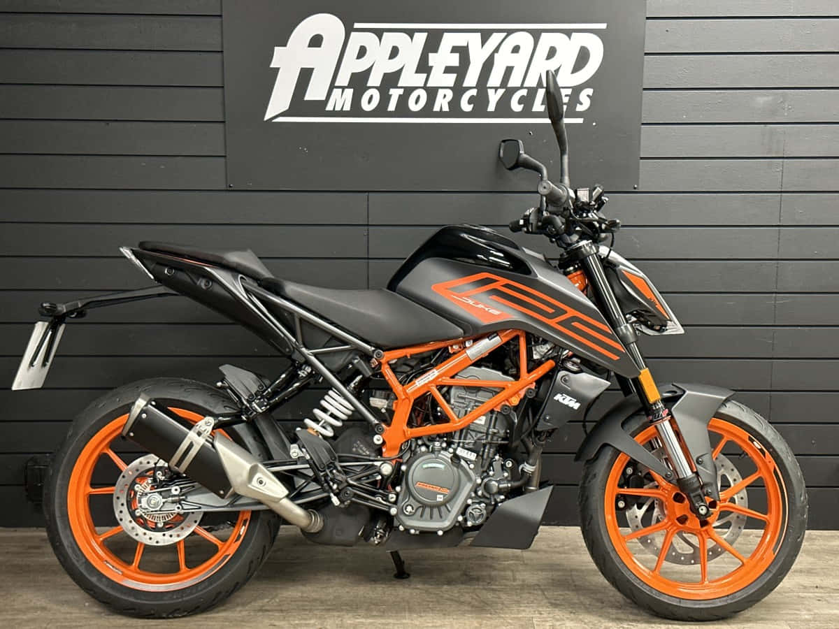 停靠在黑色牆前的KTM Duke 125機車 壁紙 — 一架時尚的黑橙色KTM電單車醒目地陳列在陳列室中，背景是深灰色條紋牆和'Appleyard Motorcycles'標誌。