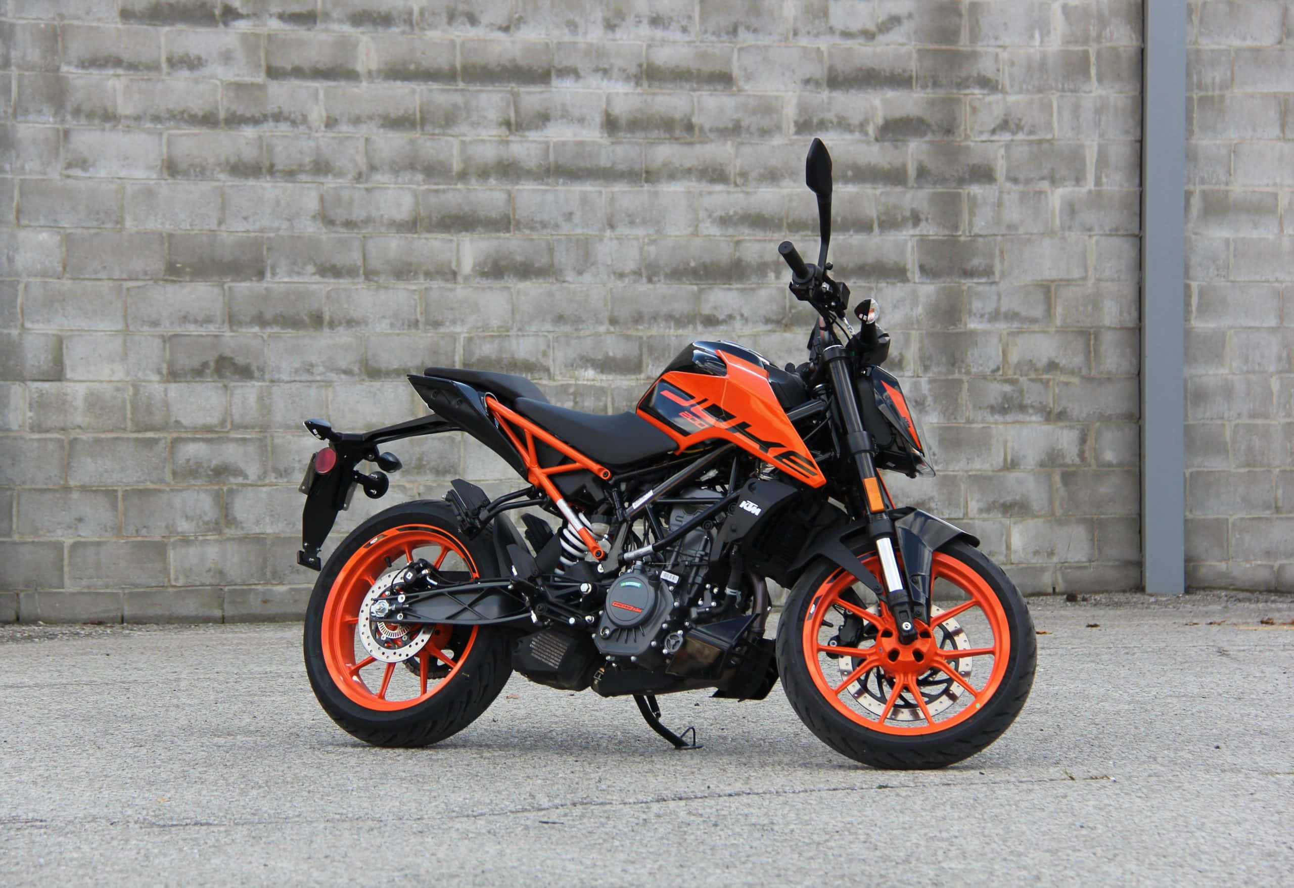 KTM Duke 200摩托車對磚牆的壯觀畫面 壁紙 — 一架搶眼的橙色和黑色KTM電單車停泊在風化的灰色磚牆前，地面是混凝土。