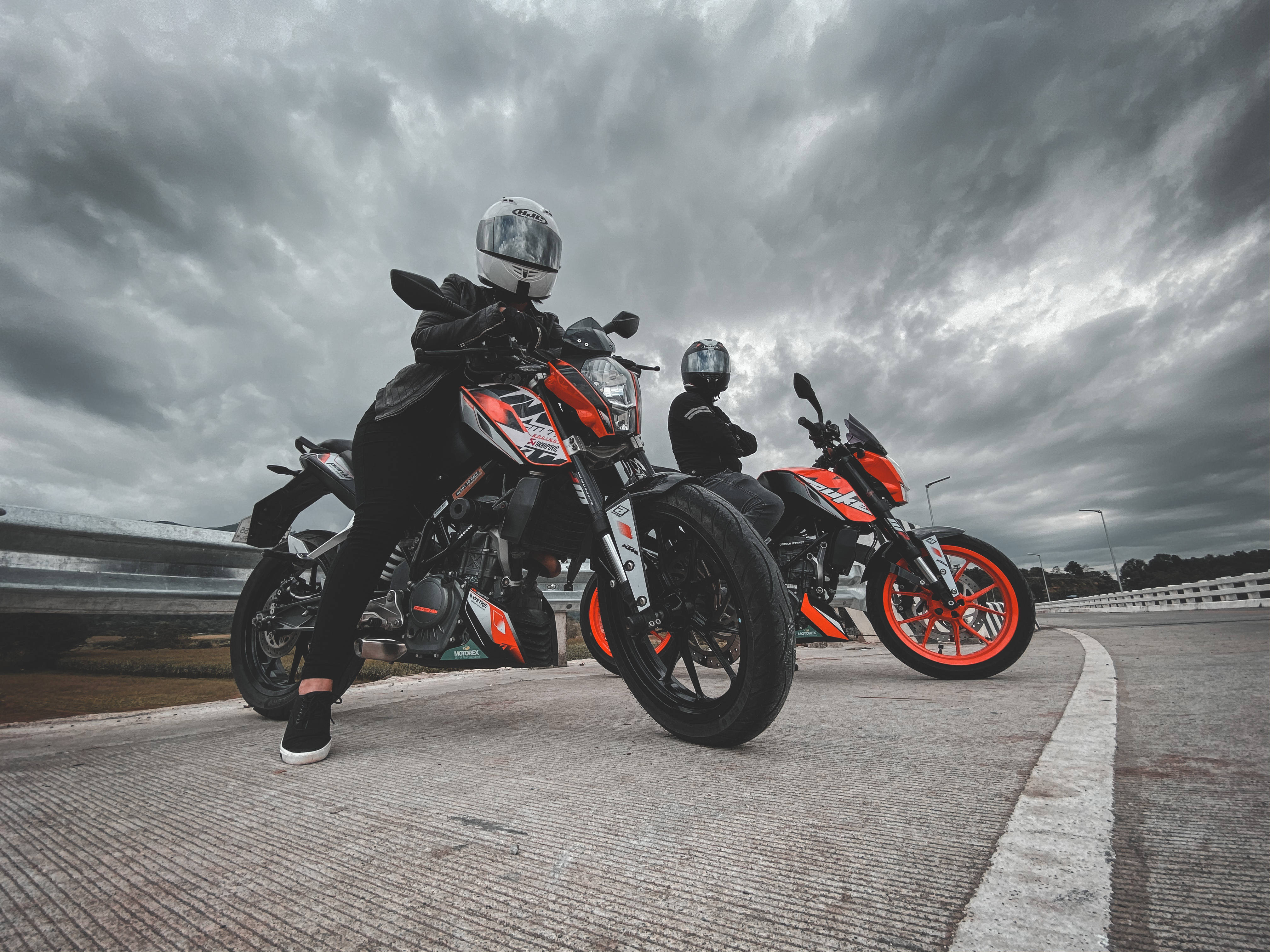 兩人坐於 KTM Duke 200 上 壁紙 — 兩名全副裝備的電單車手在戲劇性的多雲天空下，自信地擺姿勢在橙色和黑色的跑車上。