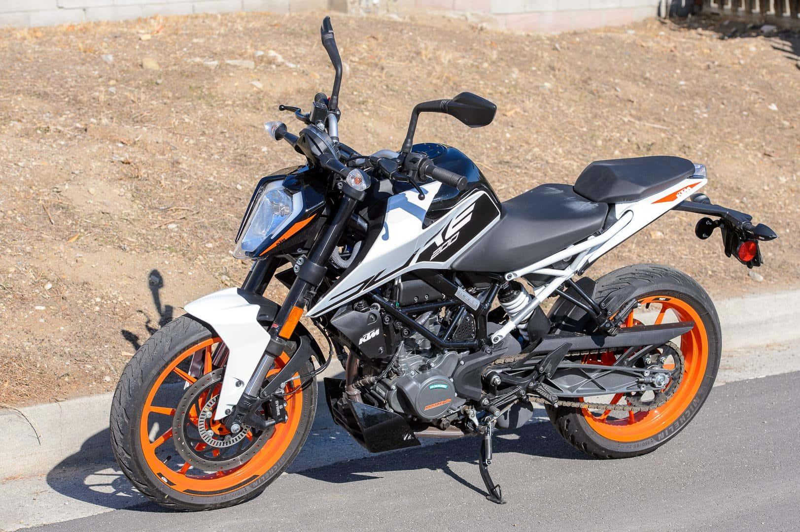 白黑配色 KTM Duke 200 機車圖片 壁紙 — 一輛時尚的KTM電單車，配備橙色車輪和白色點綴，在明亮的陽光下停泊在瀝青路面上。