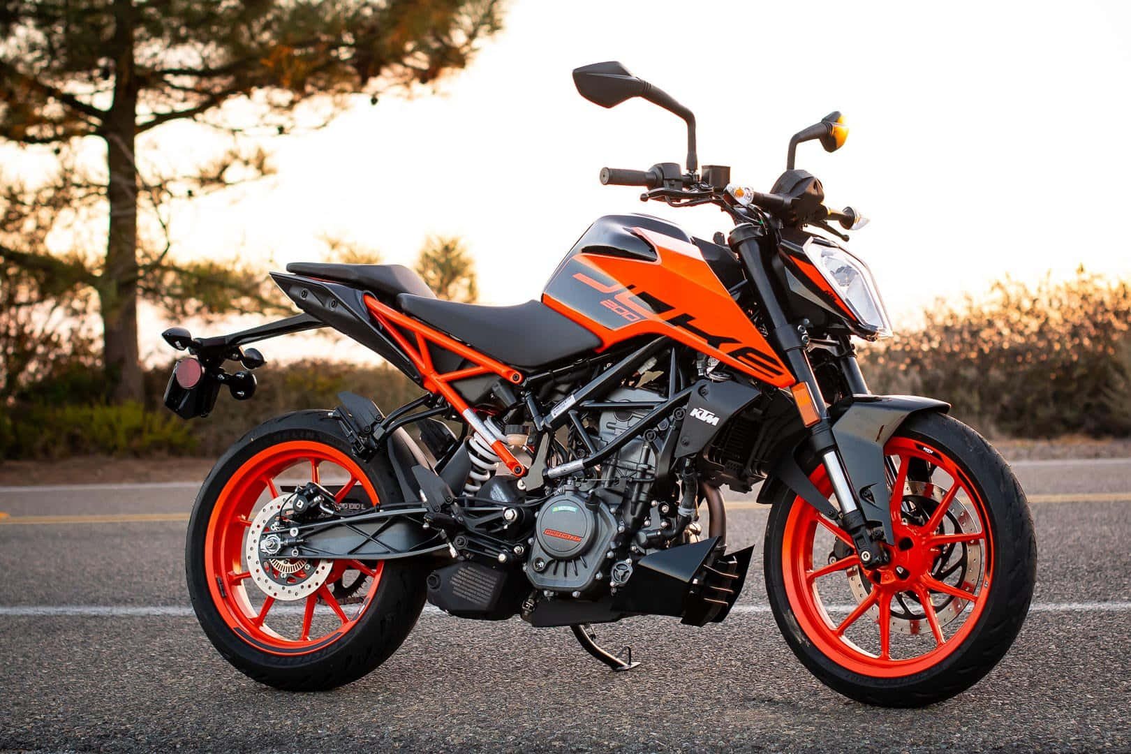 KTM Duke 200摩托車與夕陽天空 壁紙 — 一輛鮮豔的橙色和黑色KTM電單車在黃金時段停泊在瀝青路上，背景有樹木。