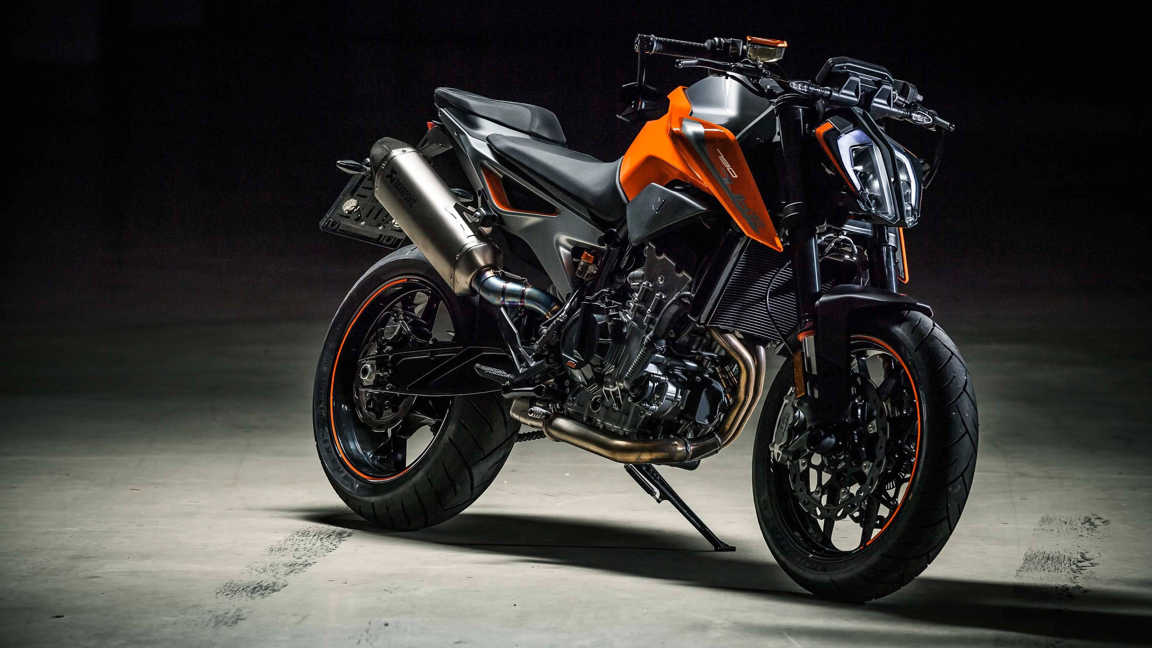 KTM Duke 390 聚光燈效果 壁紙 — 一架時尚的黑色和橙色運動電單車，帶有鍍鉻排氣管和橙色輪圈點綴，在戲劇性的工作室燈光下立於混凝土地板上。