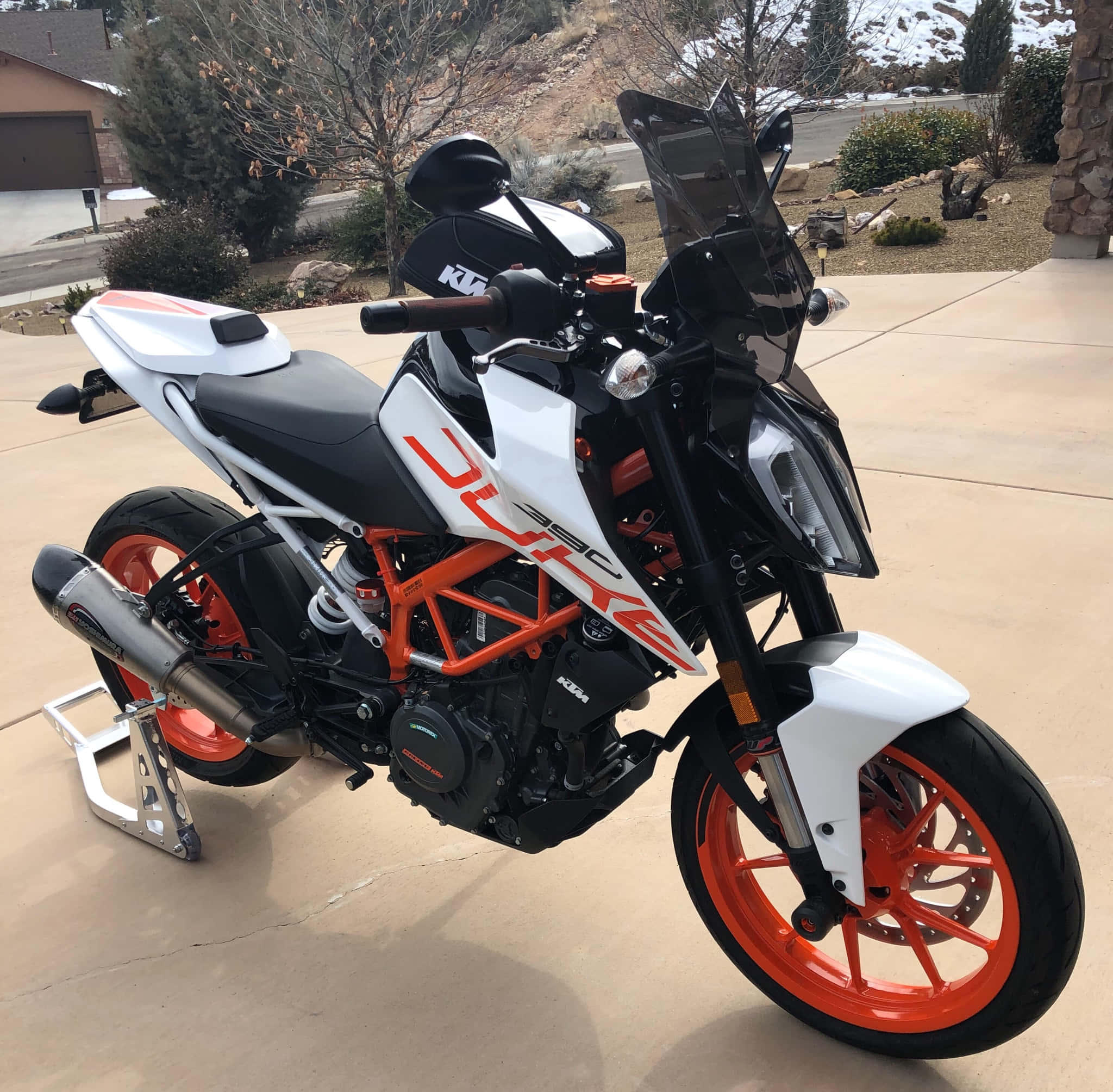 享受騎乘 KTM Duke 390 的刺激感 壁紙 — 一輛白色和橙色的KTM Duke 390電單車停放在混凝土車道上，背景是白雪覆蓋的山脈和光禿禿的樹木。