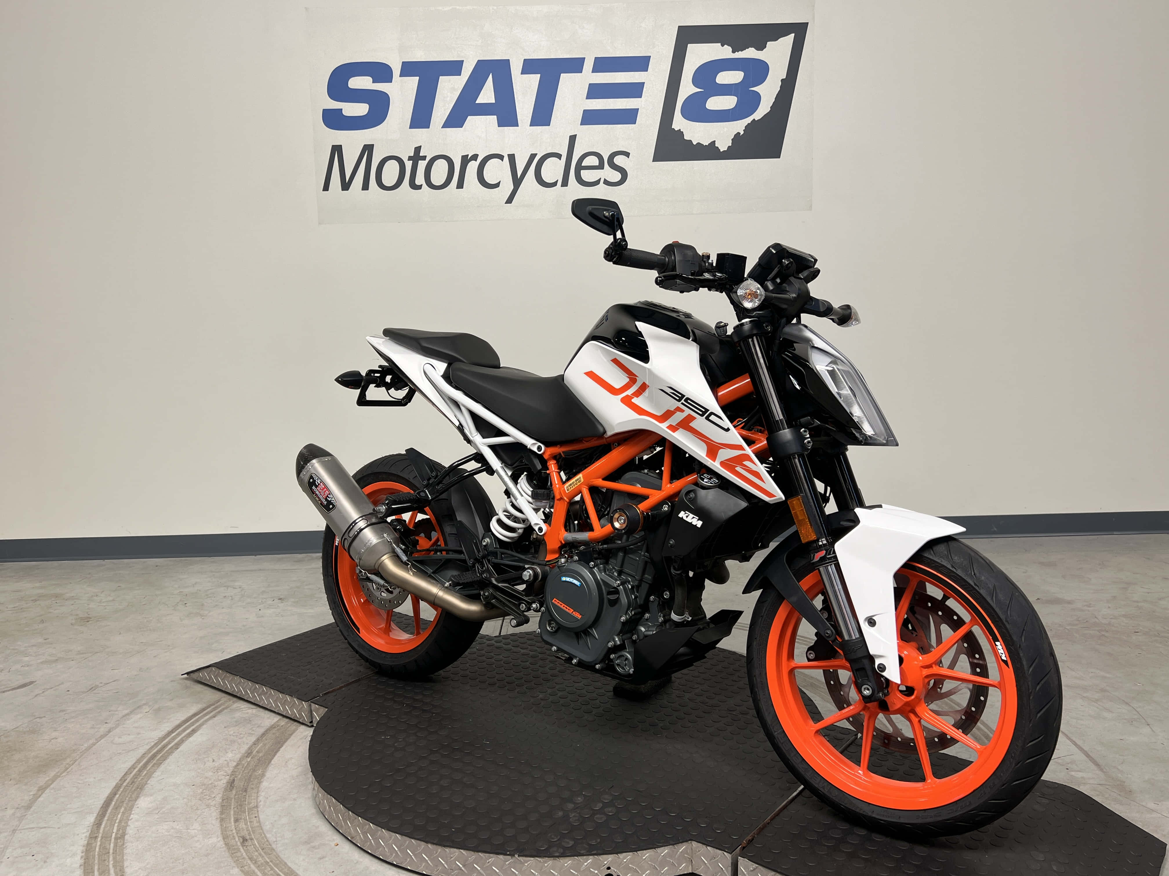 KTM Duke 390 於 State 8 Motorcycles 店前展示 壁紙 — 一輛時尚的KTM Duke電單車，帶有橙色和白色點綴，停放在展廳的黑色平台上，牆上掛有State 8 Motorcycles標誌。