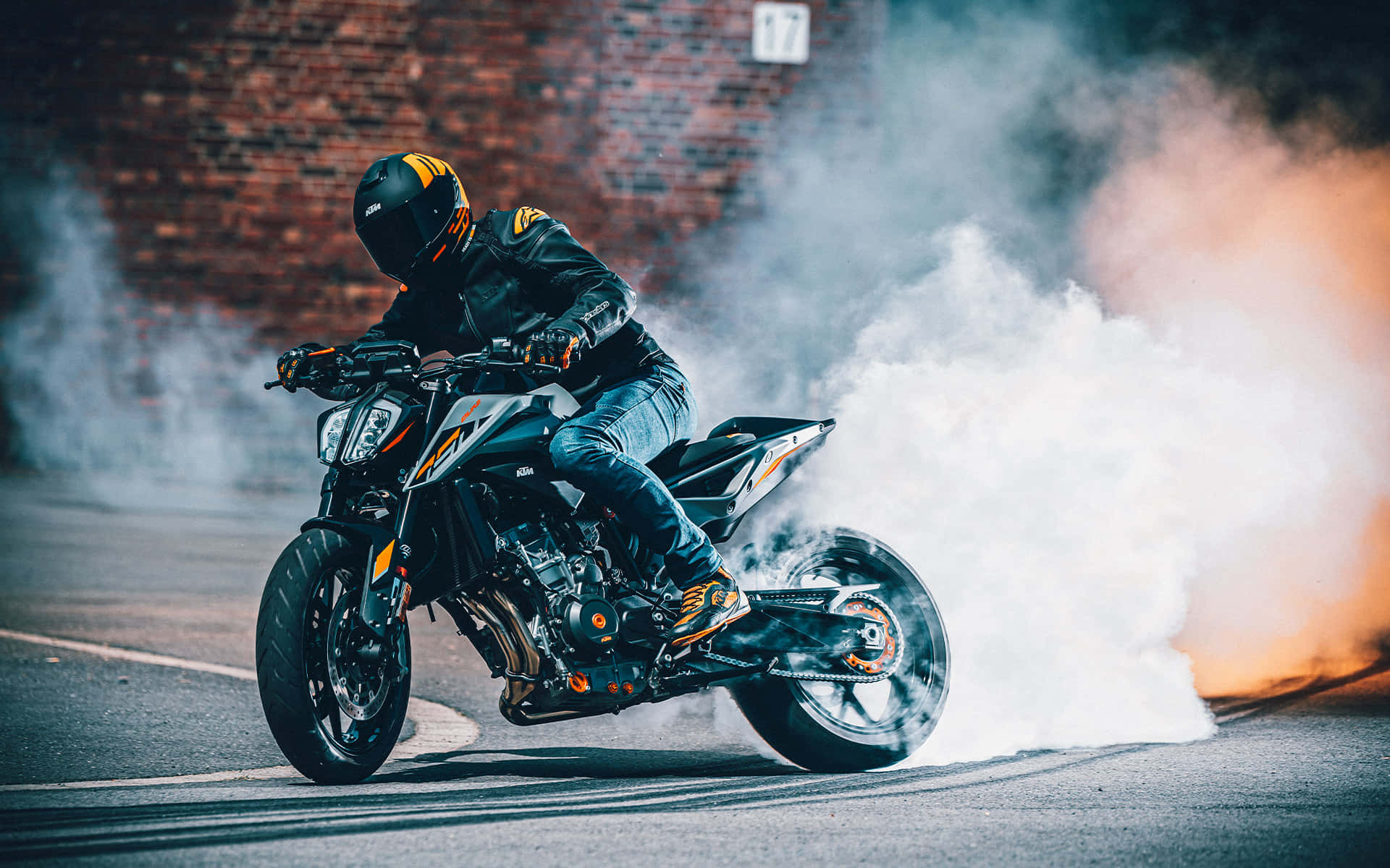 KTM Duke 790冒煙畫面 壁紙 — 一名身穿黑色裝備的電單車手在街上進行燒胎，後輪冒出濃煙，背景是磚牆。