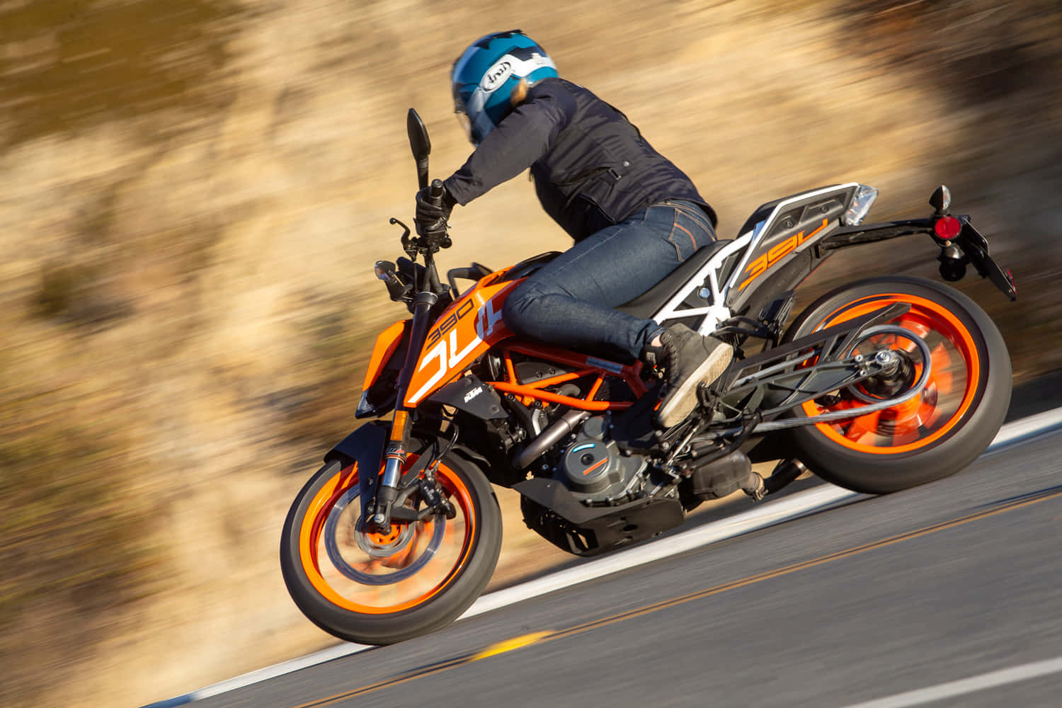 KTM Duke，準備出發 壁紙 — 一名騎士在陽光普照的道路上，騎著鮮豔的橙色KTM電單車高速行駛，傾斜入彎，背景是模糊的泥土色調。