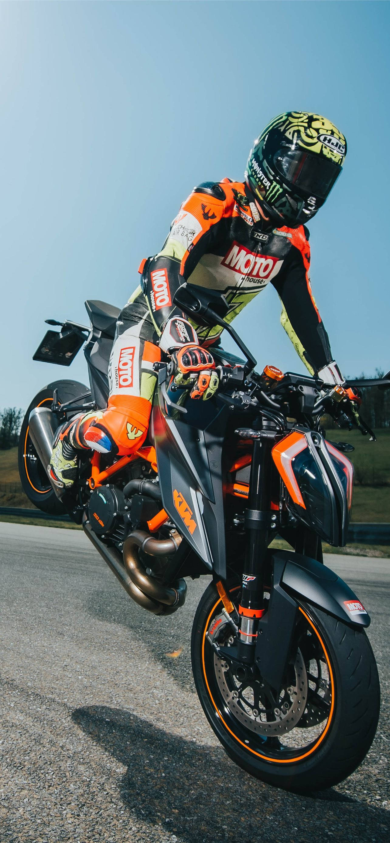 專業賽車手 KTM iPhone 壁紙 — 一名身穿橙色裝備的電單車手在晴朗的藍天下，駕駛跑車電單車表演前輪離地。