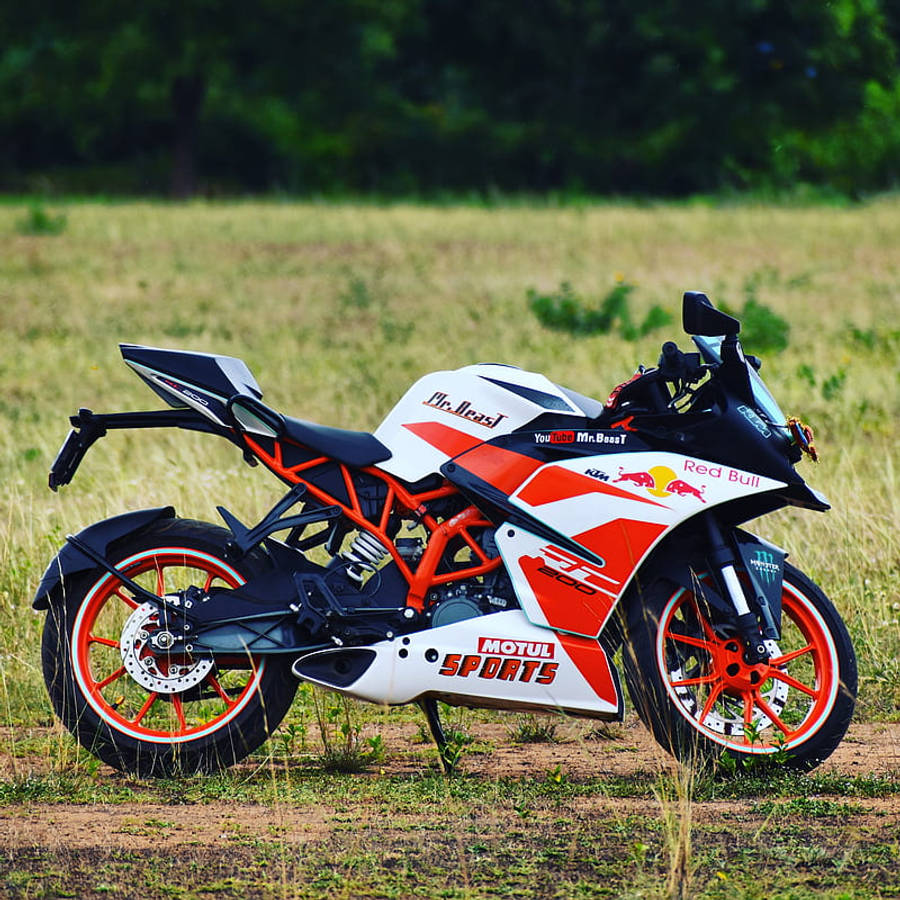 賽車塗裝KTM RC 200 壁紙 — 一架白色、橙色和黑色的KTM RC390電單車，帶有Red Bull和MrBeast貼紙，停泊在陽光明媚的草地上。