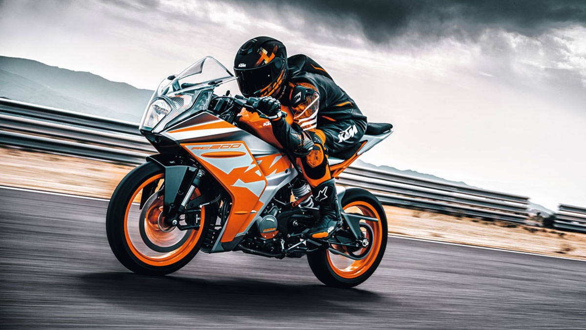 騎士駕駛KTM RC 200 壁紙 — 一名身穿黑色和橙色裝備的車手在暴風雨天空下的賽道上，騎著鮮豔的橙色和銀色KTM電單車傾斜入彎。