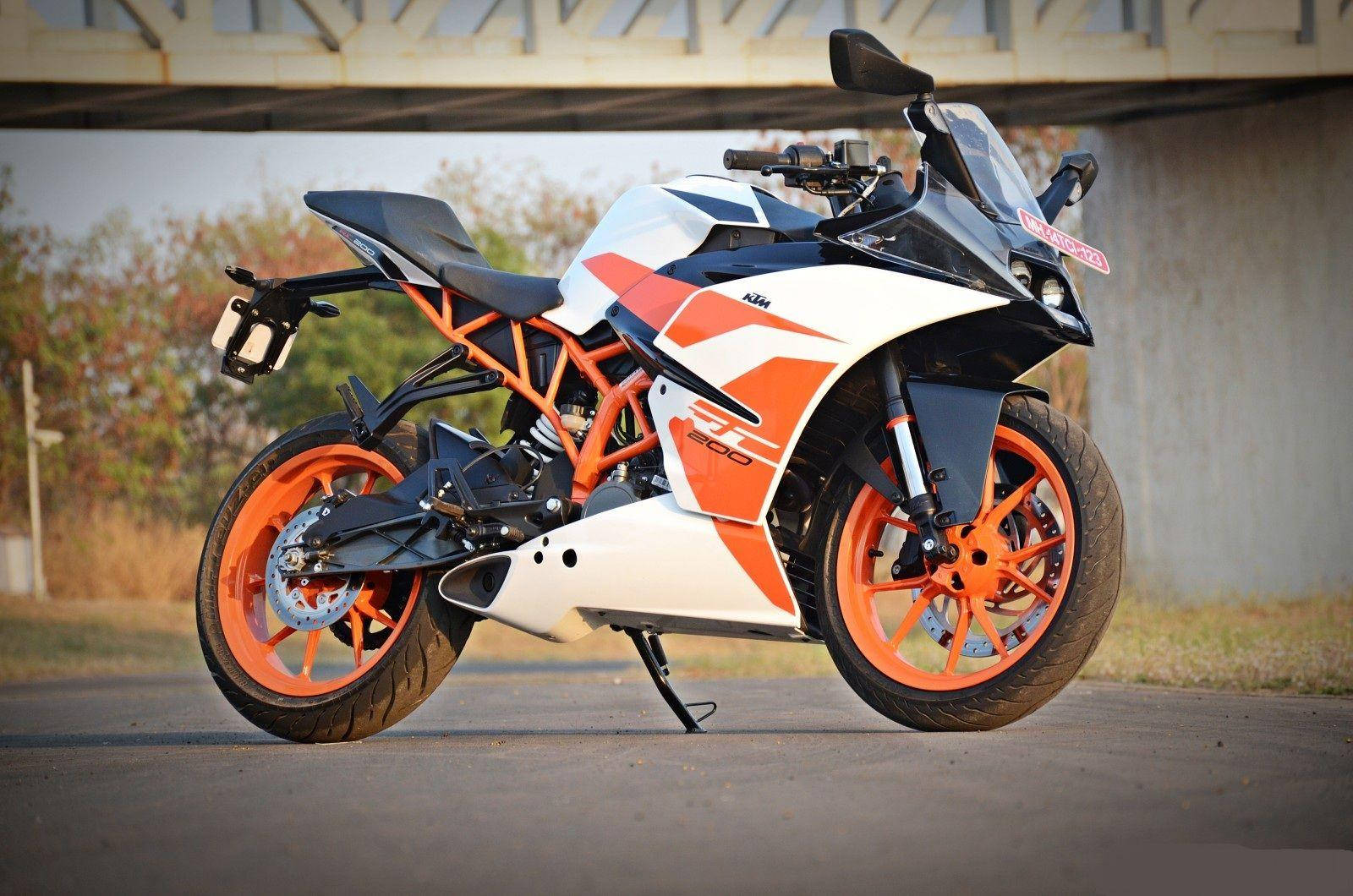 城市風景中的KTM RC 200 壁紙 — 一架時尚的白色和橙色KTM運動電單車停泊在混凝土橋下，背景有秋天的樹木。