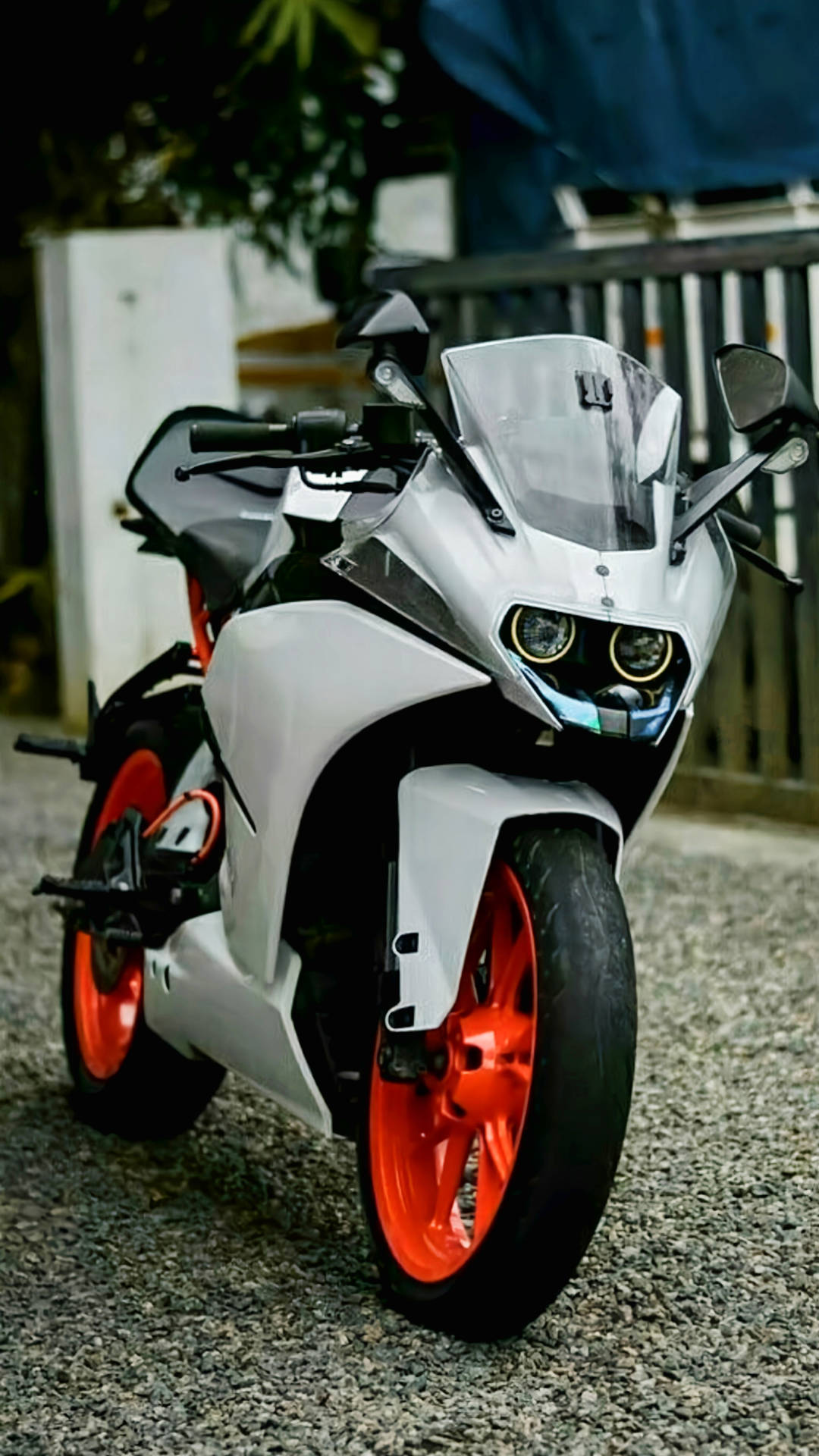 清爽白色KTM RC 200 壁紙 — 一架時尚的白色運動電單車，配以鮮艷橙色輪胎，停泊在圍欄附近的碎石地上。